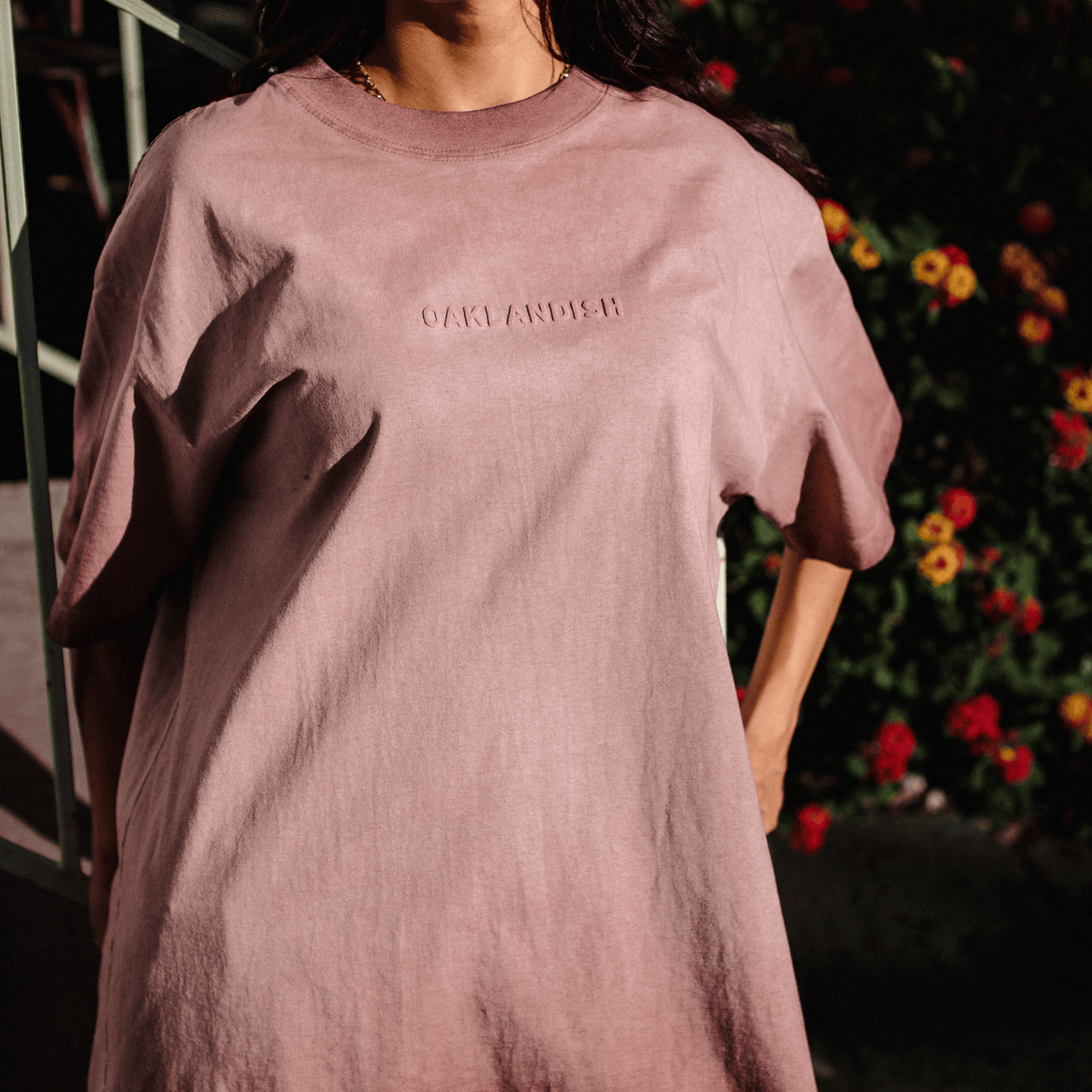 A person wears a dusty rose-colored Oaklandish Classic Logo Tee featuring the word “OAKLANDISH” in puff print across the chest, set against a backdrop of dark foliage with hints of yellow and red flowers.