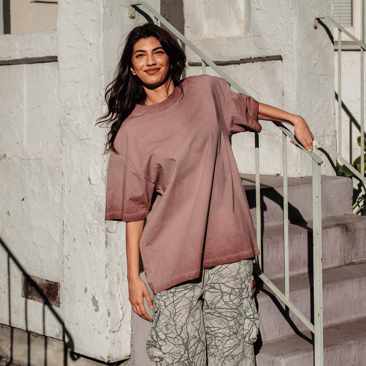 A woman with long dark hair and a nose ring stands on outdoor stairs, smiling in the sunlight while wearing an Oaklandish Classic Logo Tee in muted mauve and light-colored cargo pants with a dark, branch-like pattern, leaning against a white stucco wall.