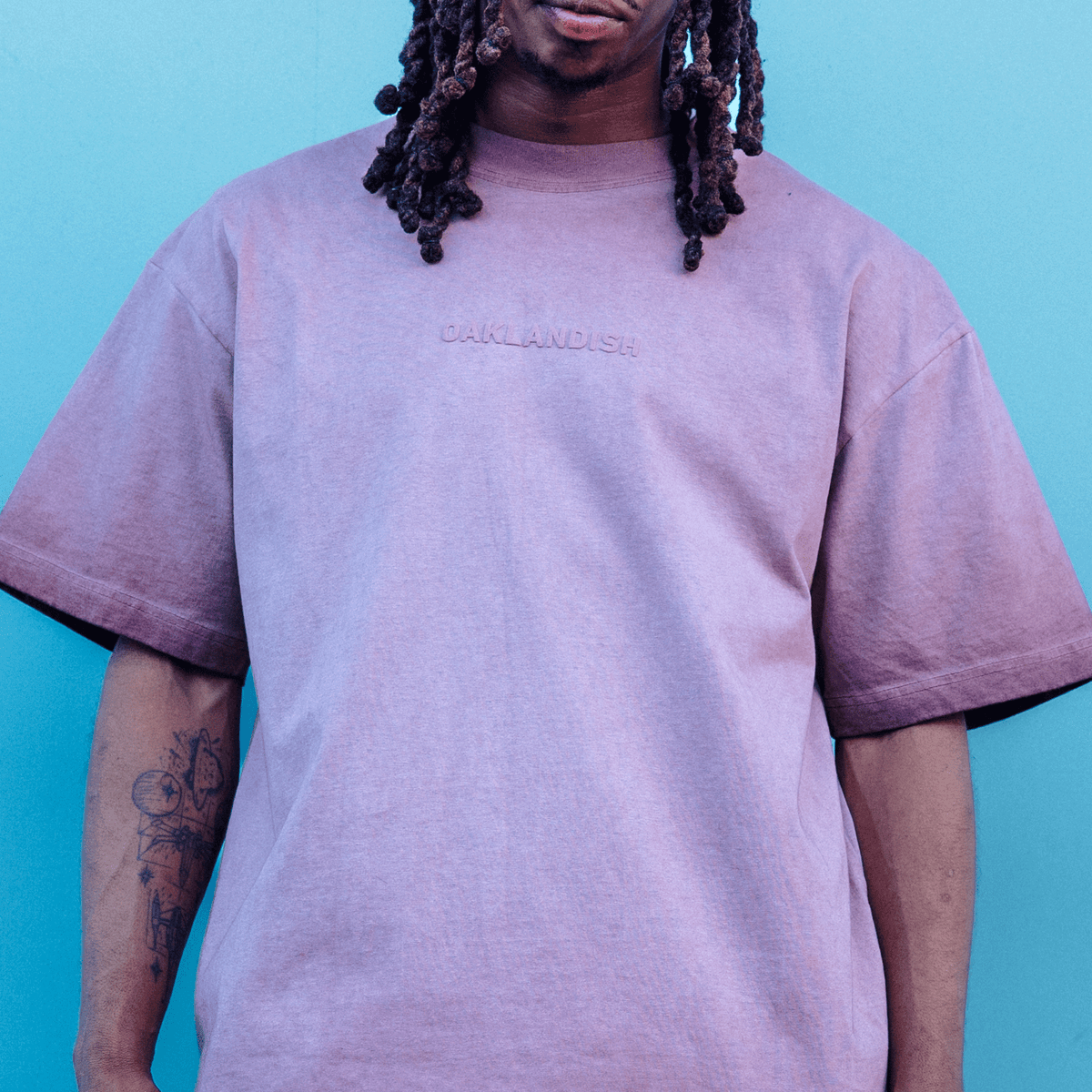 A person with dreadlocks and a visible forearm tattoo wears a mauve Oaklandish Classic Logo Tee with “OAKLANDISH” in puff print across the chest against a solid, bright blue background.