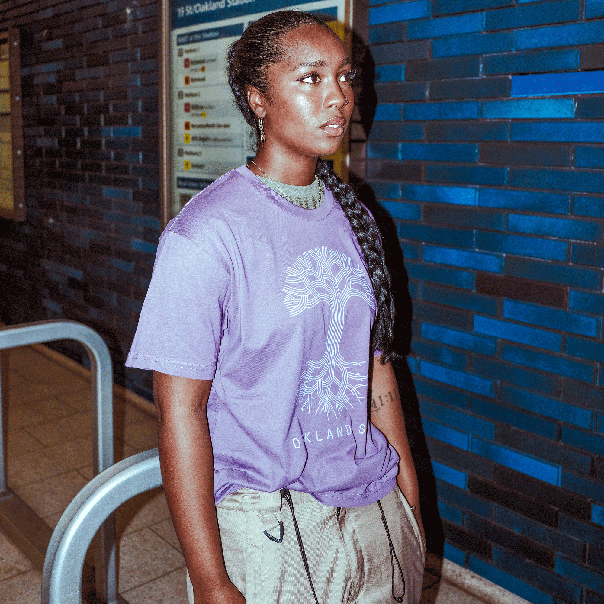 An individual with long, braided hair stands indoors near a blue-tiled wall, wearing the Oaklandish Classic Logo Tee by Oaklandish and tan pants. They face slightly sideways with a neutral expression; a subway sign appears in the background.