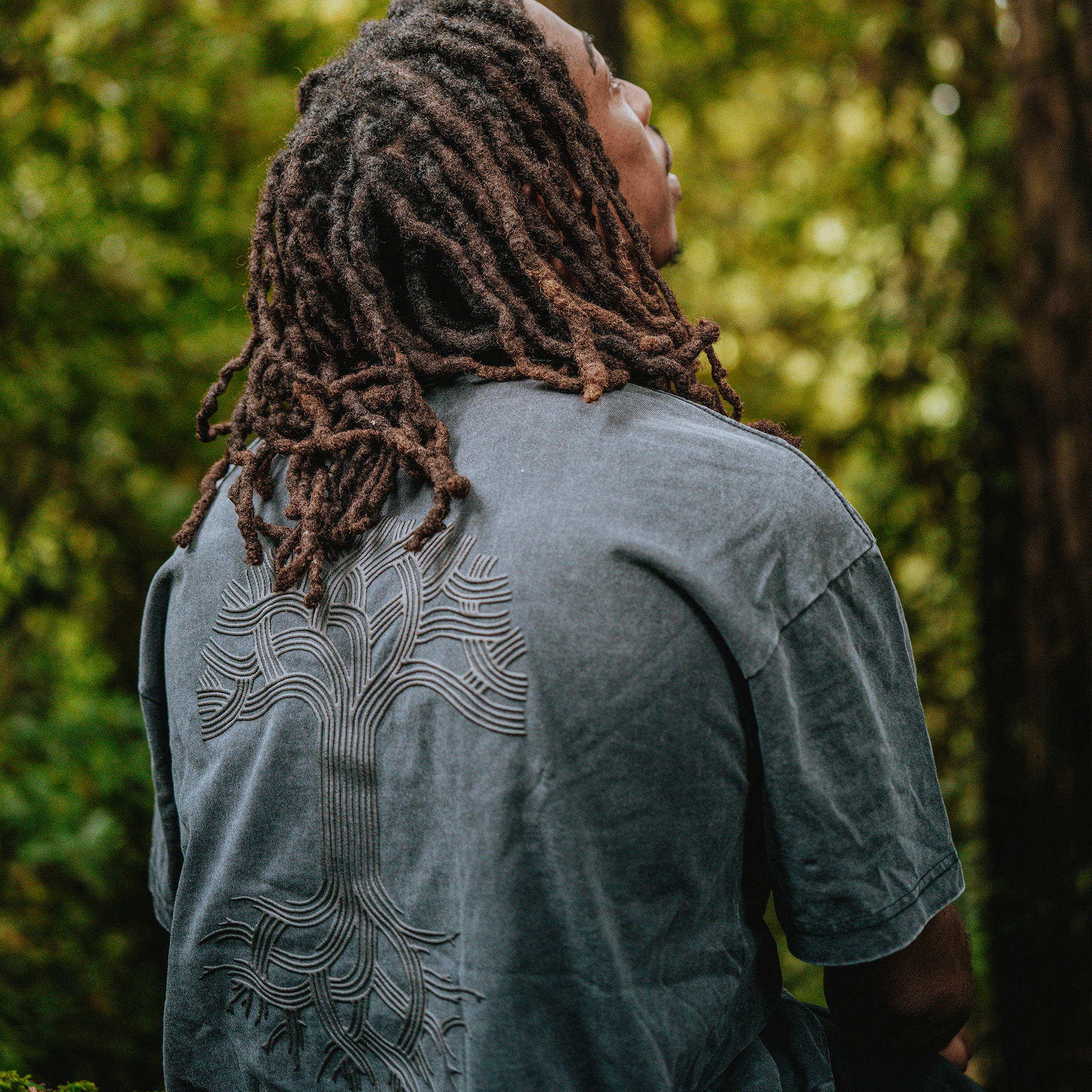 A person with long, brown dreadlocks is seen from behind wearing the Oaklandish Classic Logo Tee by Oaklandish, a faded gray men's cotton t-shirt featuring the brand’s iconic tree logo, while standing outdoors amid lush greenery.