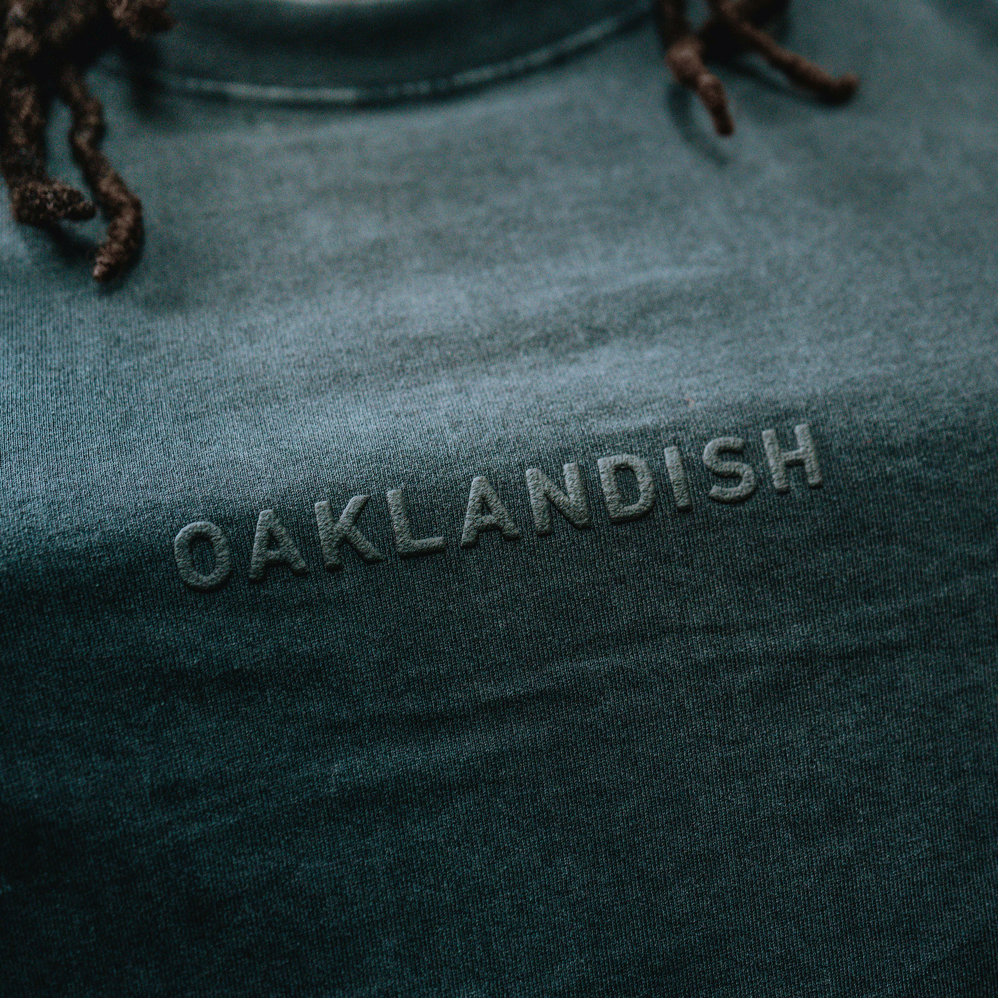 Close-up of the Oaklandish Classic Logo Tee by Oaklandish, a dark green men’s cotton t-shirt featuring "OAKLANDISH" embossed on the chest. Dark braids accent its soft, slightly worn texture and classic fit.