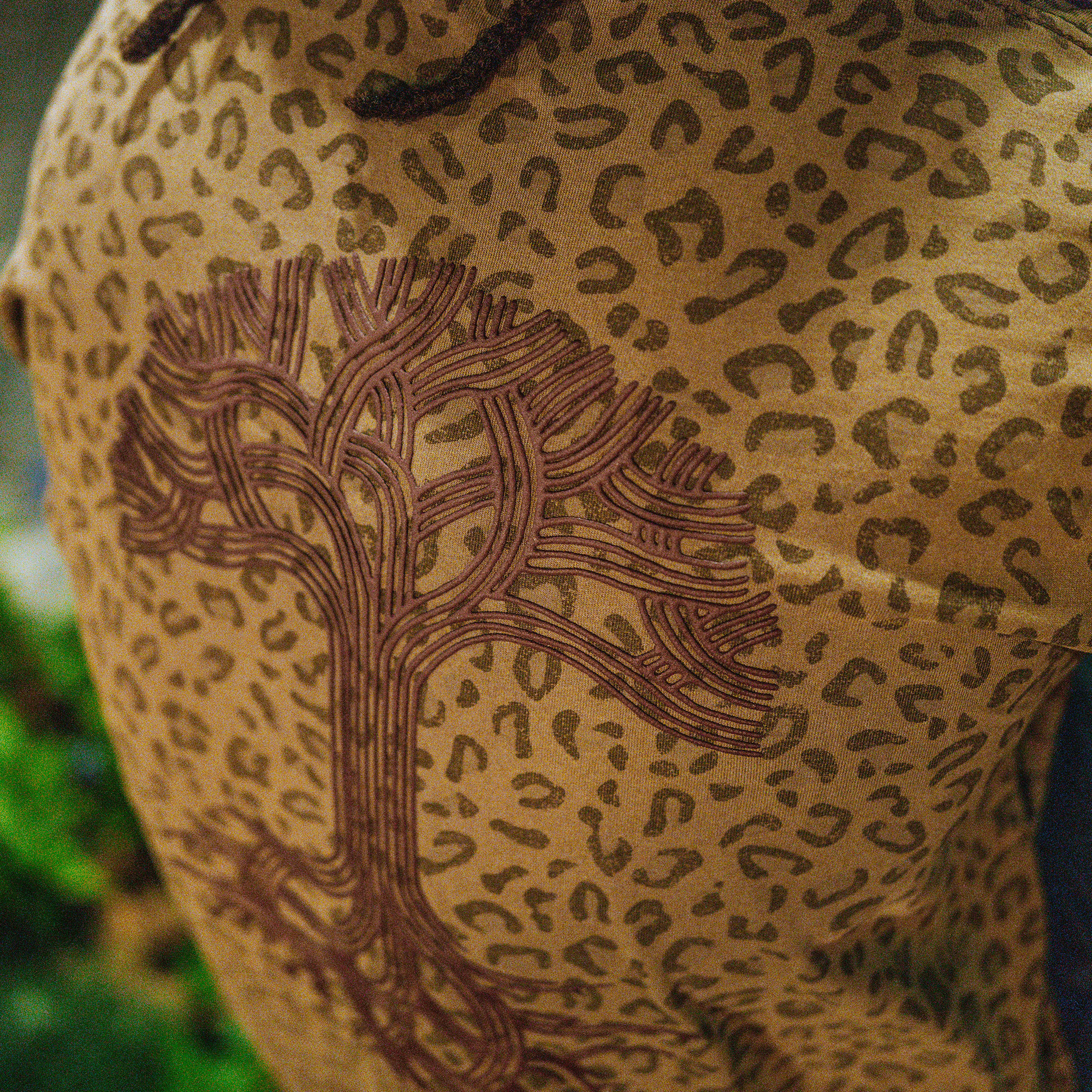 A close-up of the Oaklandish Classic Logo Tee by Oaklandish, showing a large brown tree logo on tan cotton with dark leopard-print spots and blurred greenery in the lower left.