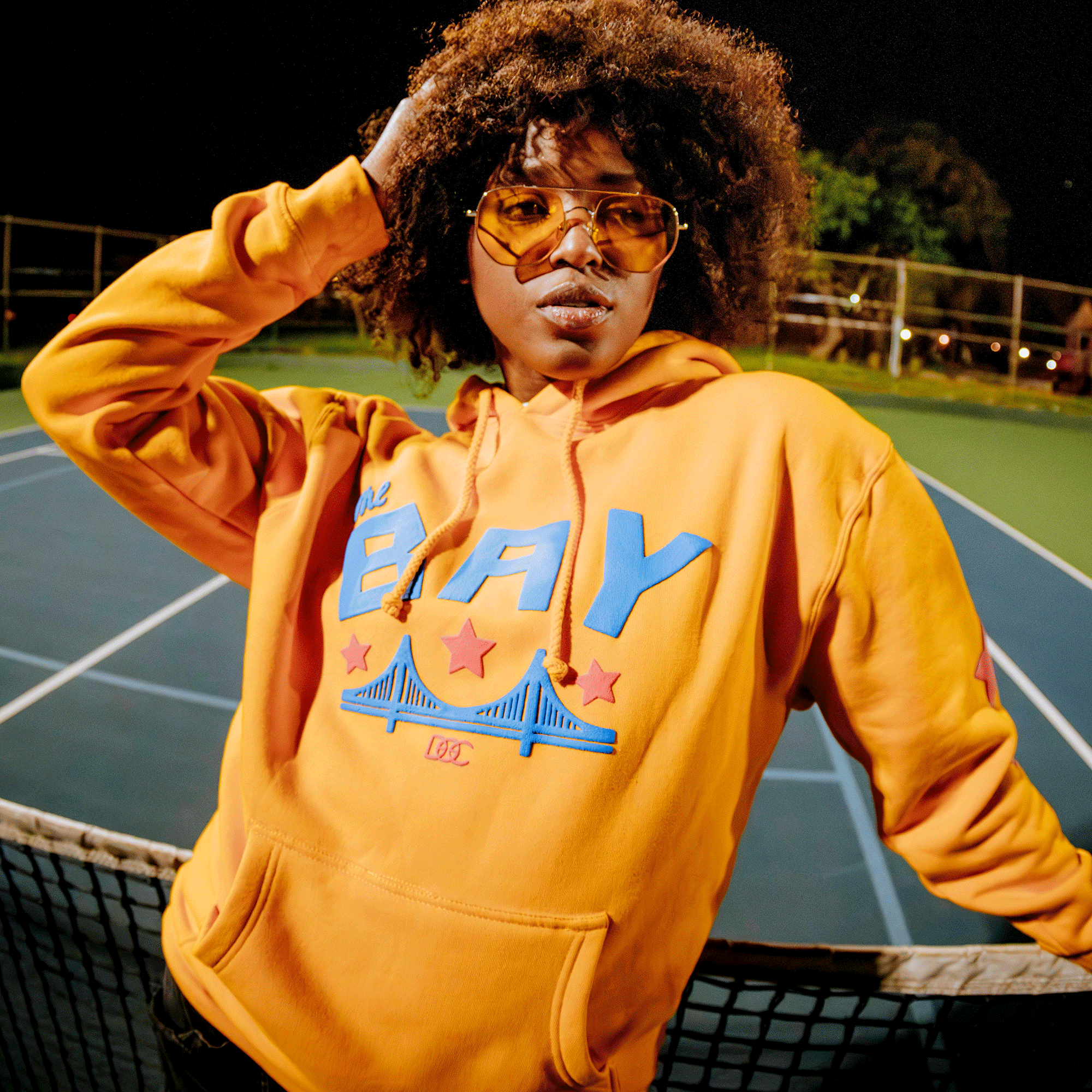 On a night-lit tennis court, a person with curly hair, yellow-tinted sunglasses, and an orange Bay Bridge Hoodie by DOC from Oaklandish stands confidently with one hand on their head and the other on the net, radiating Bay Area global culture.