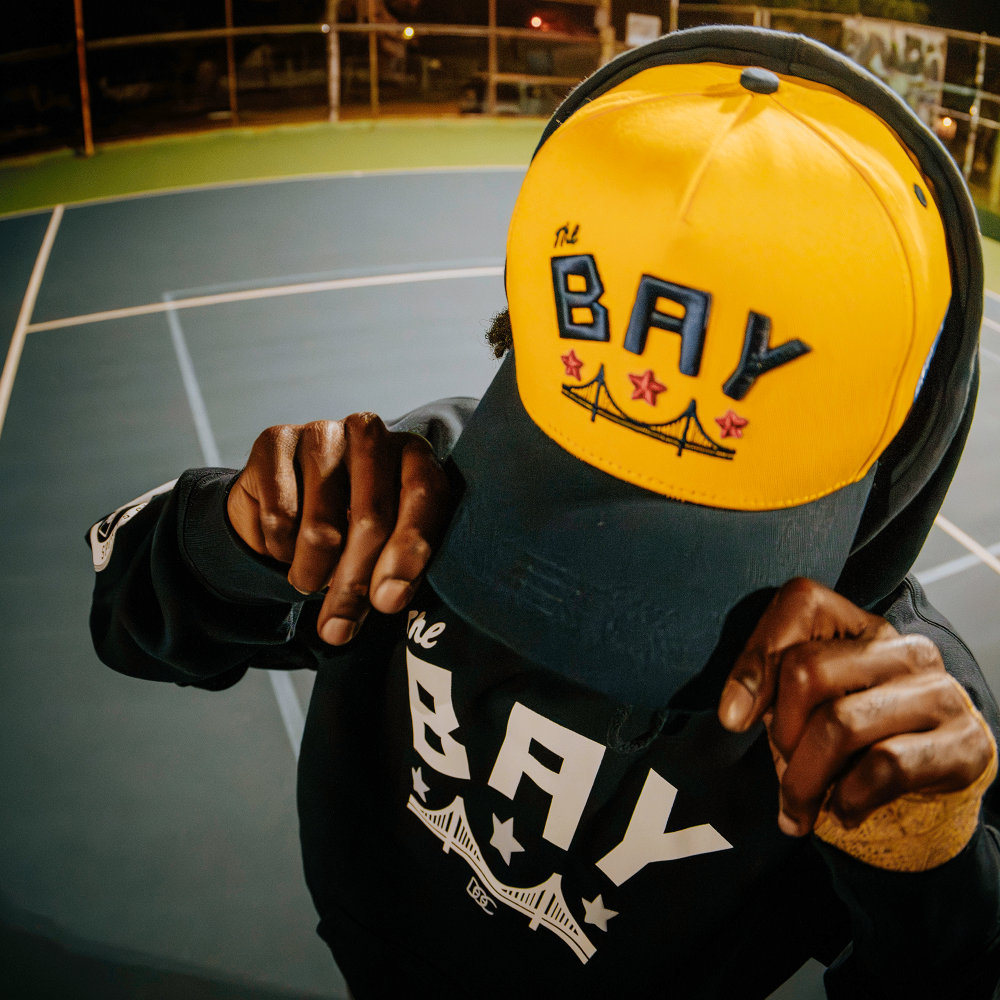 On the tennis court, someone adjusts the yellow and navy Bay Bridge Snapback by DOC from Oaklandish, featuring "THE BAY" and a bridge design. Wearing a matching hoodie, their hands and hat are the focus as they stand over the court lines.