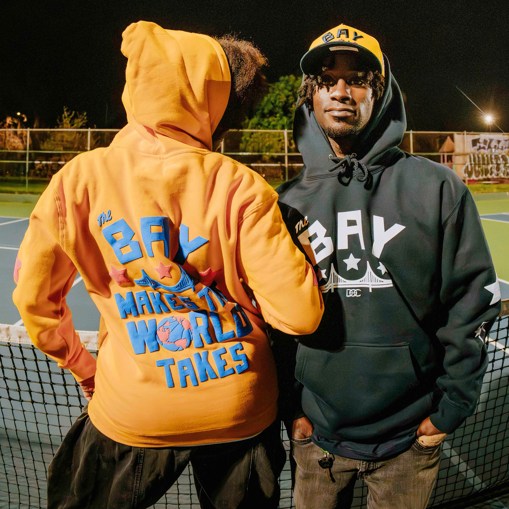 Two people stand on a tennis court at night in Oaklandish Bay Bridge Hoodies by DOC, channeling Bay Area style. One faces forward in black and yellow; the other’s orange hoodie features bold text, a globe, and stars.