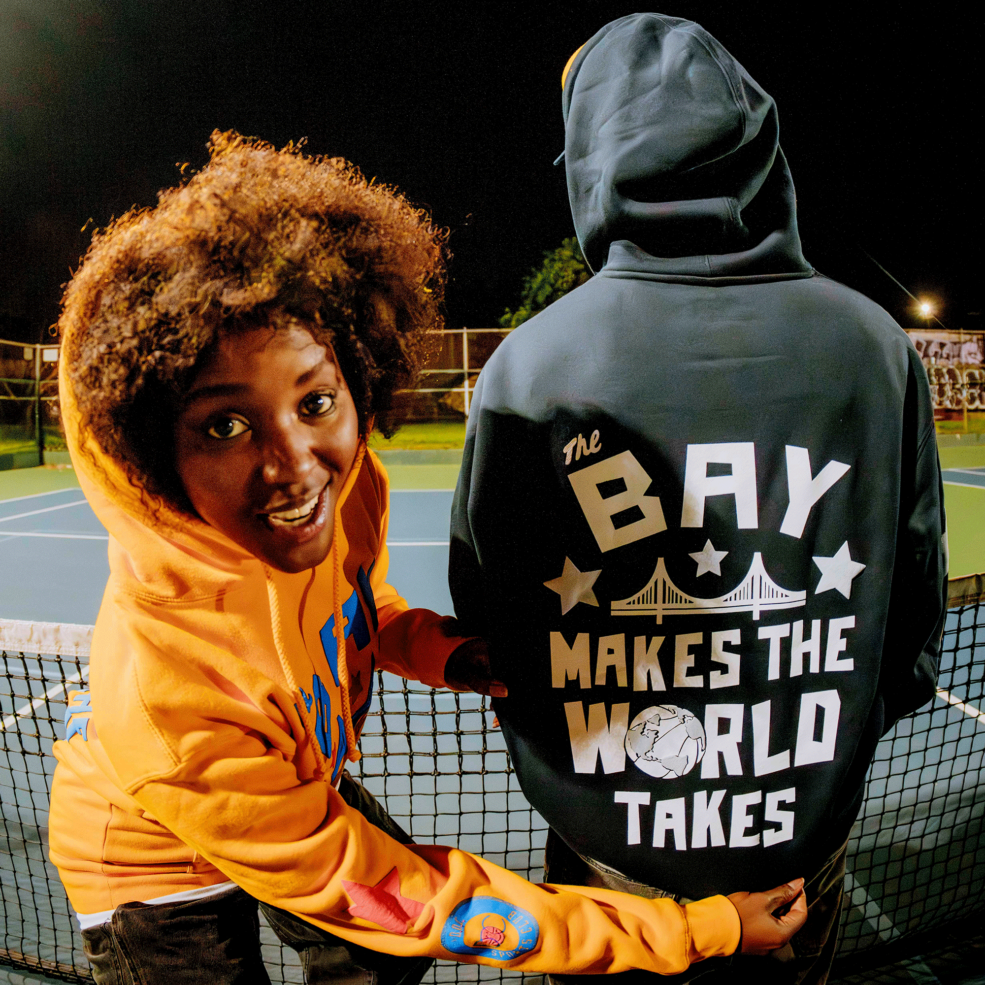 Two people are on a tennis court at night. One smiles at the camera in an orange hoodie. The other, showcasing local pride in Oaklandish’s Bay Bridge Hoodie by DOC, faces away, highlighting Bay Area-inspired style under the night sky.
