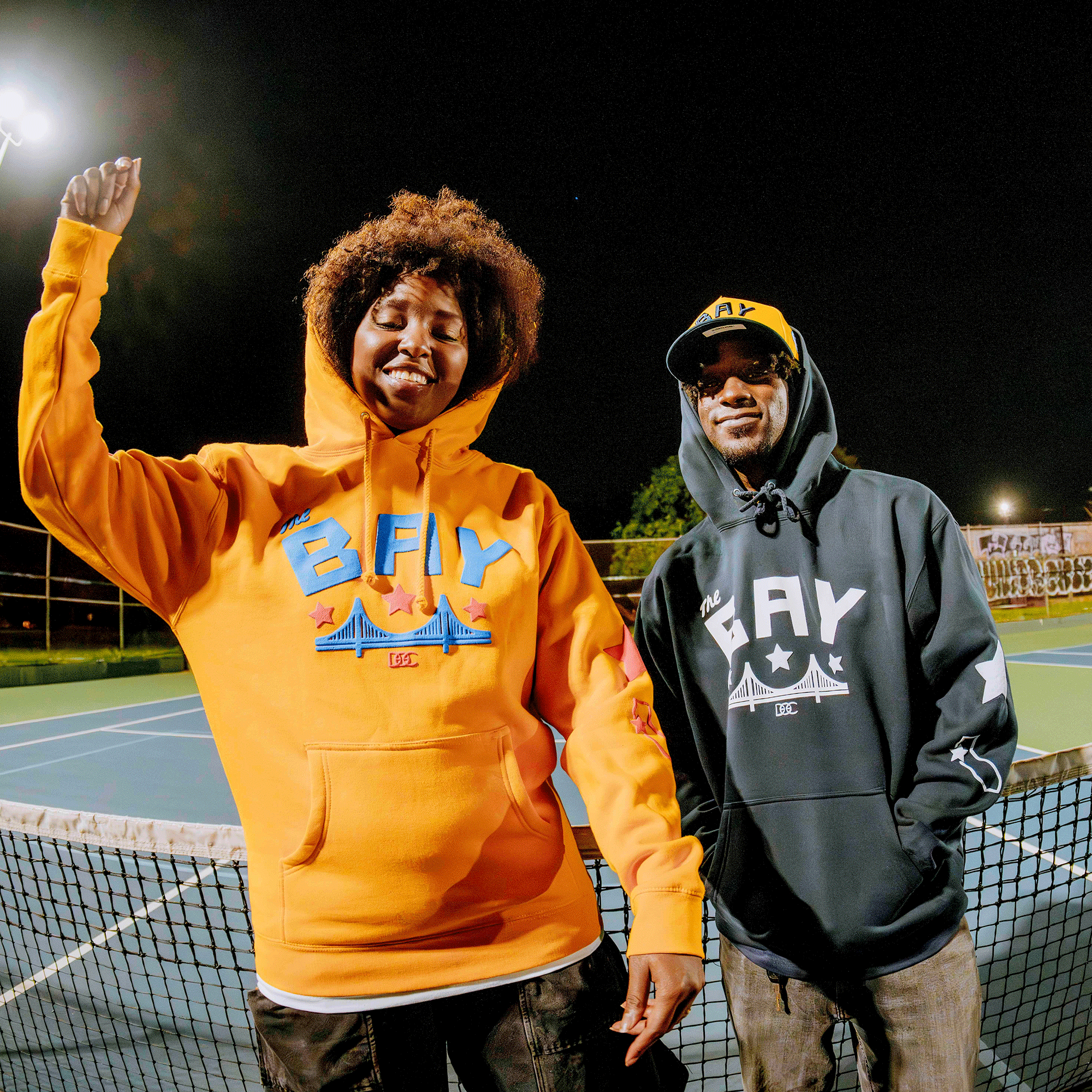 Two people stand on a tennis court at night, smiling in Bay Bridge Hoodies by DOC from Oaklandish. Sporting stylized bridge graphics and “BAY” text, they celebrate Bay Area pride. The tennis net and court are visible in the background.