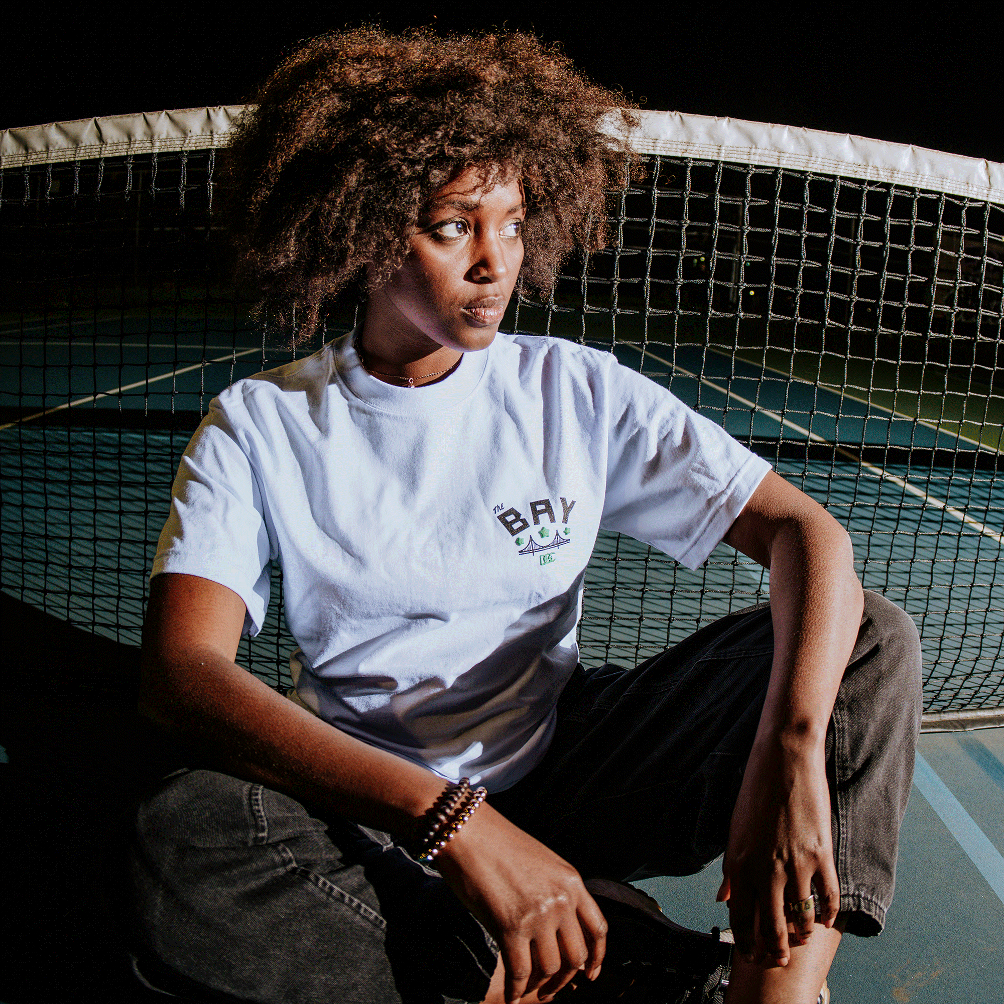 A woman with curly hair sits cross-legged on a tennis court at night, wearing the Oaklandish Bay Bridge Tee by DOC and dark jeans, gazing to the side under bright lights, channeling Bay Area culture.