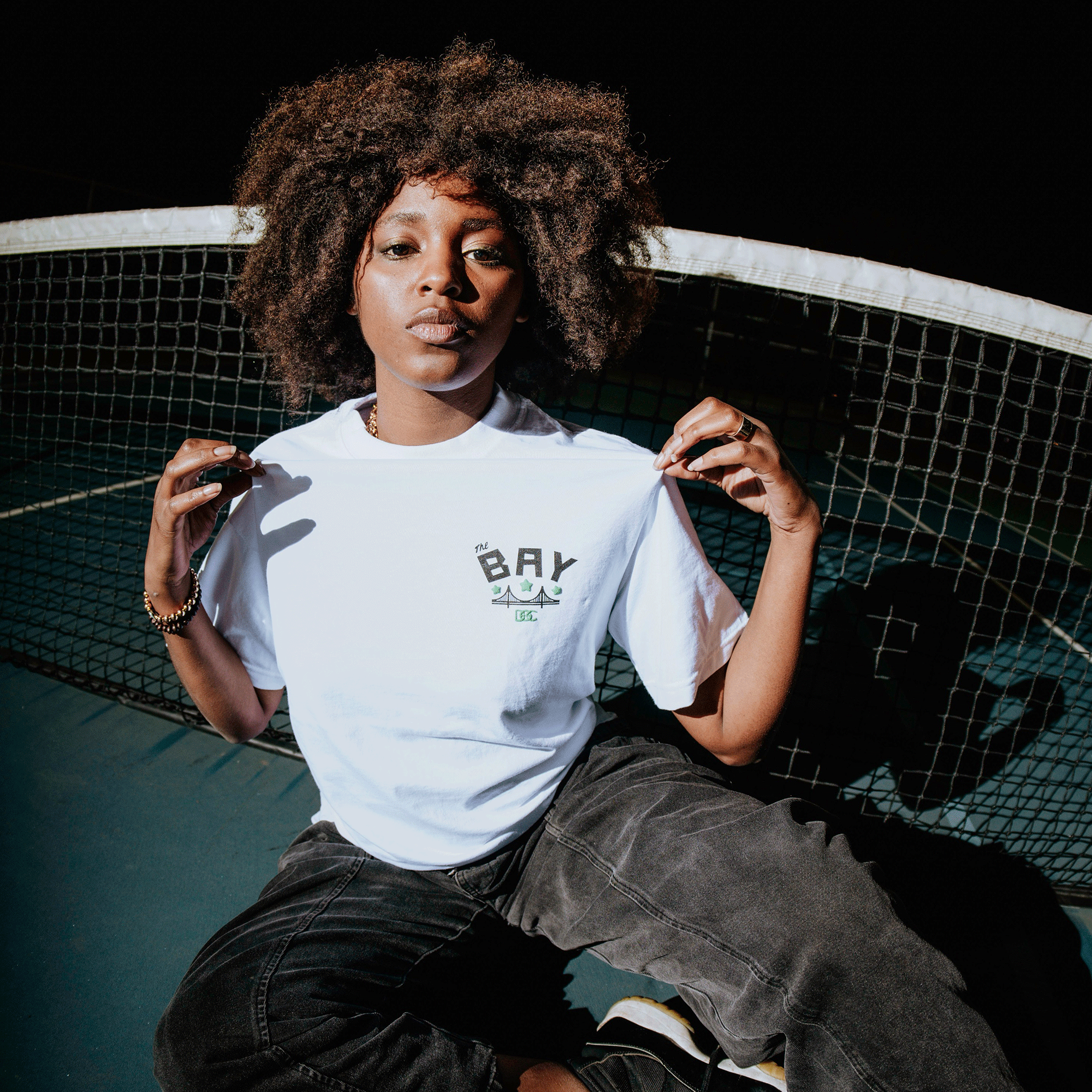 A woman with natural curly hair sits in front of a tennis net at night, confidently holding out her white Bay Bridge Tee by DOC from Oaklandish—"BAY" in bold letters—paired with dark jeans under dramatic lighting.
