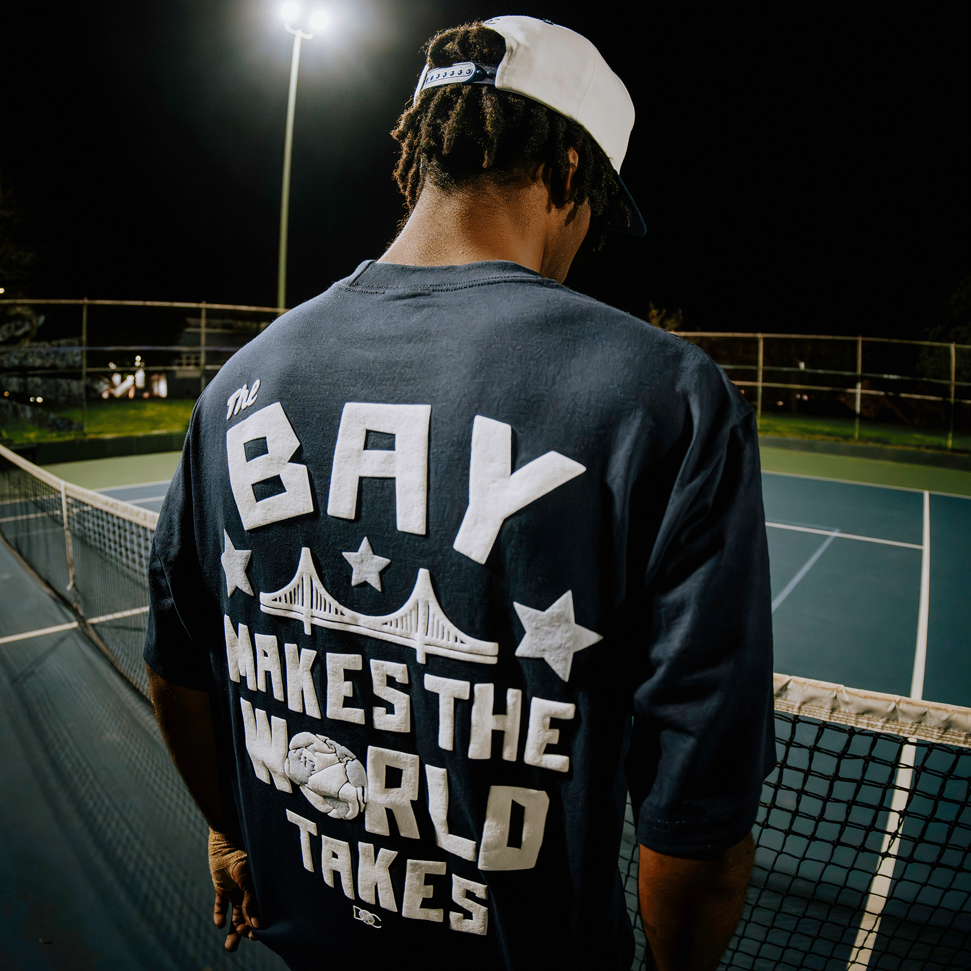 A man stands on a lit outdoor tennis court at night, wearing a backward white cap and the Bay Bridge Tee by DOC from Oaklandish, featuring “The Bay Makes The World Takes” and an illustration of a bridge and basketball.