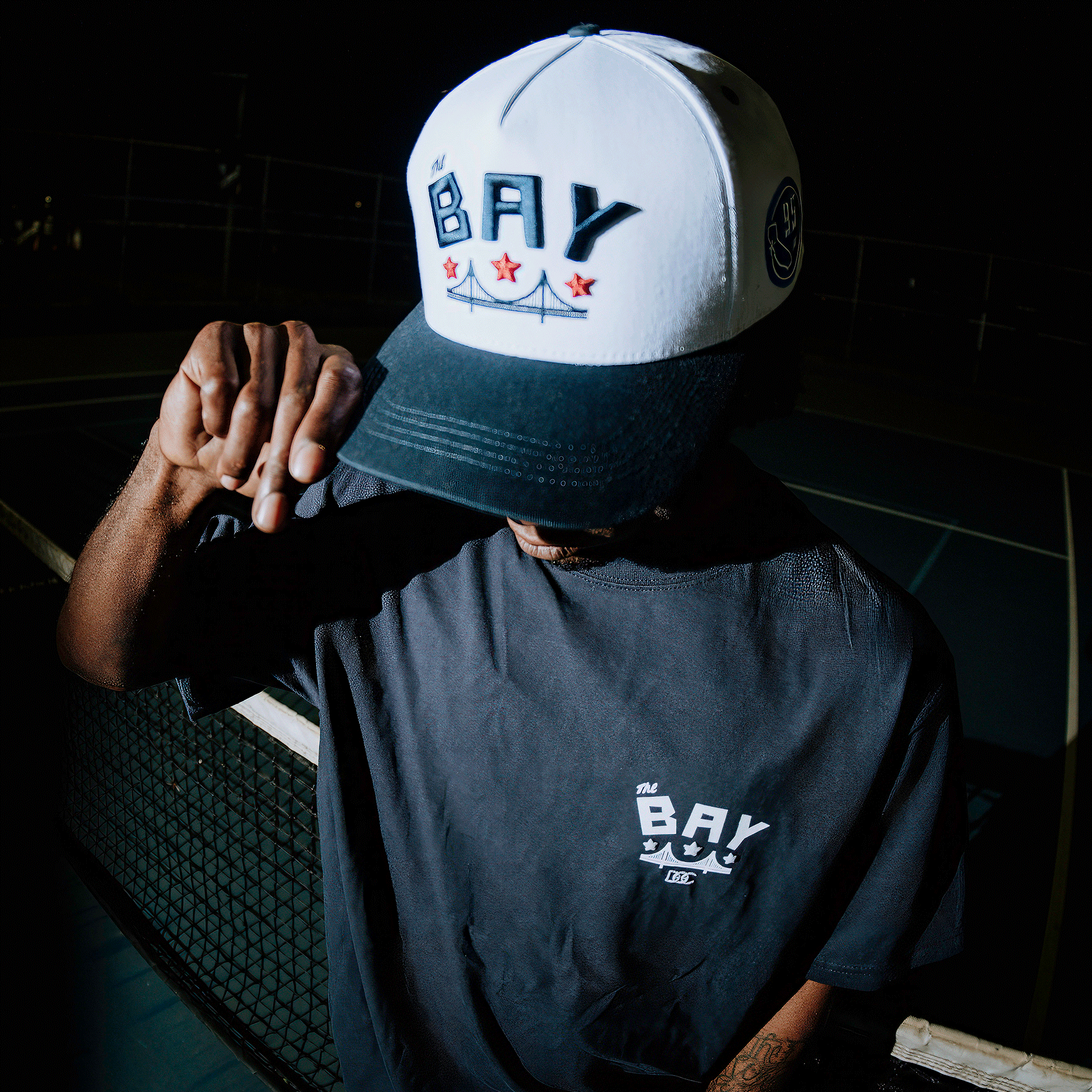 A person stands at night on a dark outdoor sports court, tilting the Oaklandish Bay Bridge Snapback by DOC to shade their face; they wear a black T-shirt and channel Bay Area culture by the chain-link fence.