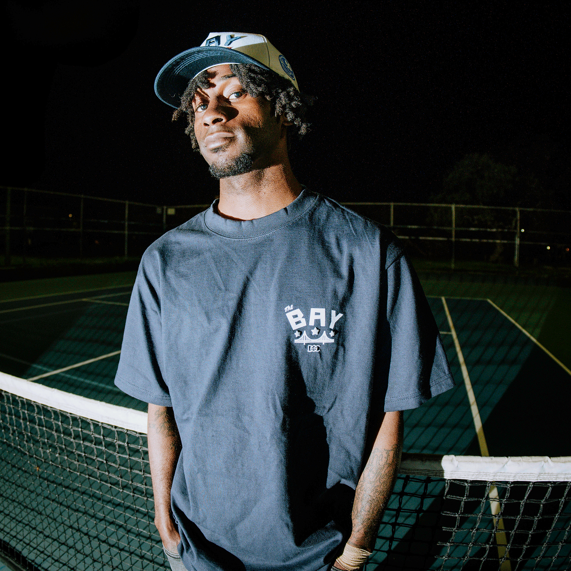A confident young man with short dreadlocks stands on an empty tennis court at night, wearing the Oaklandish Bay Bridge Tee by DOC, embodying the spirit of "The Bay Makes the World Takes.