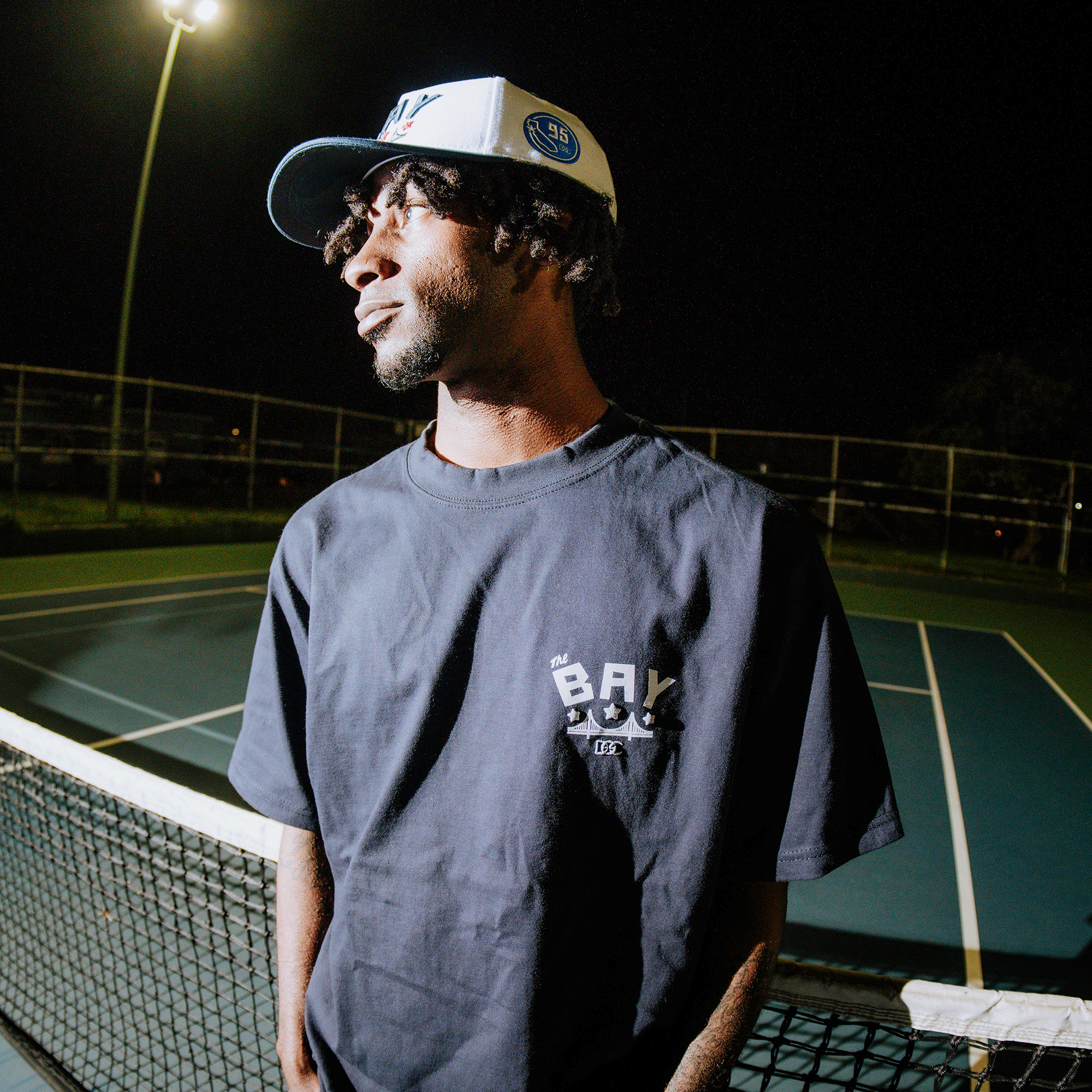 A young man in a dark Oaklandish "Bay Bridge Tee by DOC" stands by a tennis net on an outdoor court at night, under overhead lights—capturing a moody Bay Area vibe inspired by local art themes.