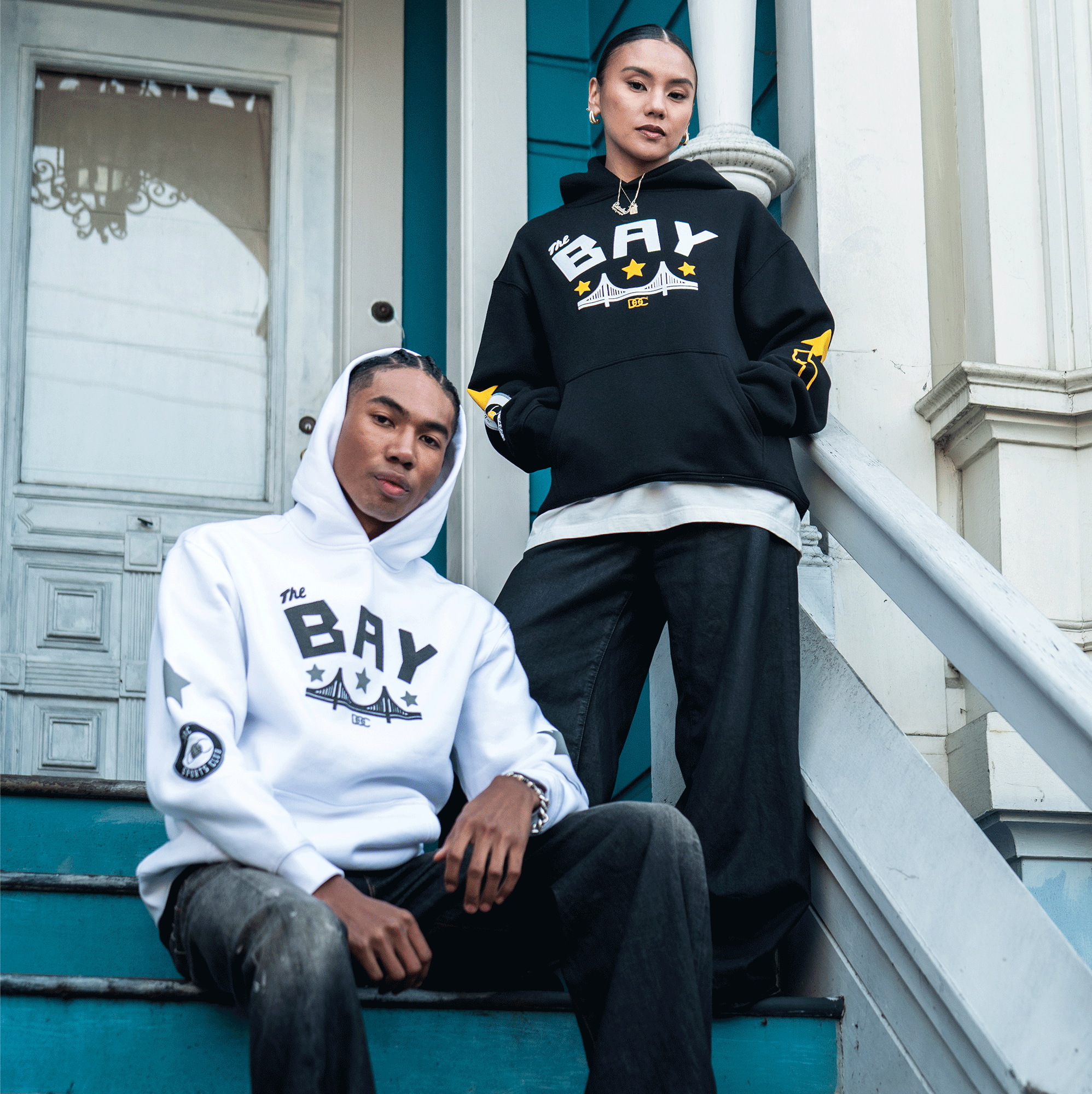 Two people pose on blue steps outside a house. One wears the Bay Bridge Hoodie By DOC V2 from Oaklandish in white; the other stands in a black men's outerwear hoodie. Both wear loose jeans and look serious, with a white door behind them.