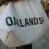 One side of tote bag as worn on person with text Oaklandish in black text on cream tote.