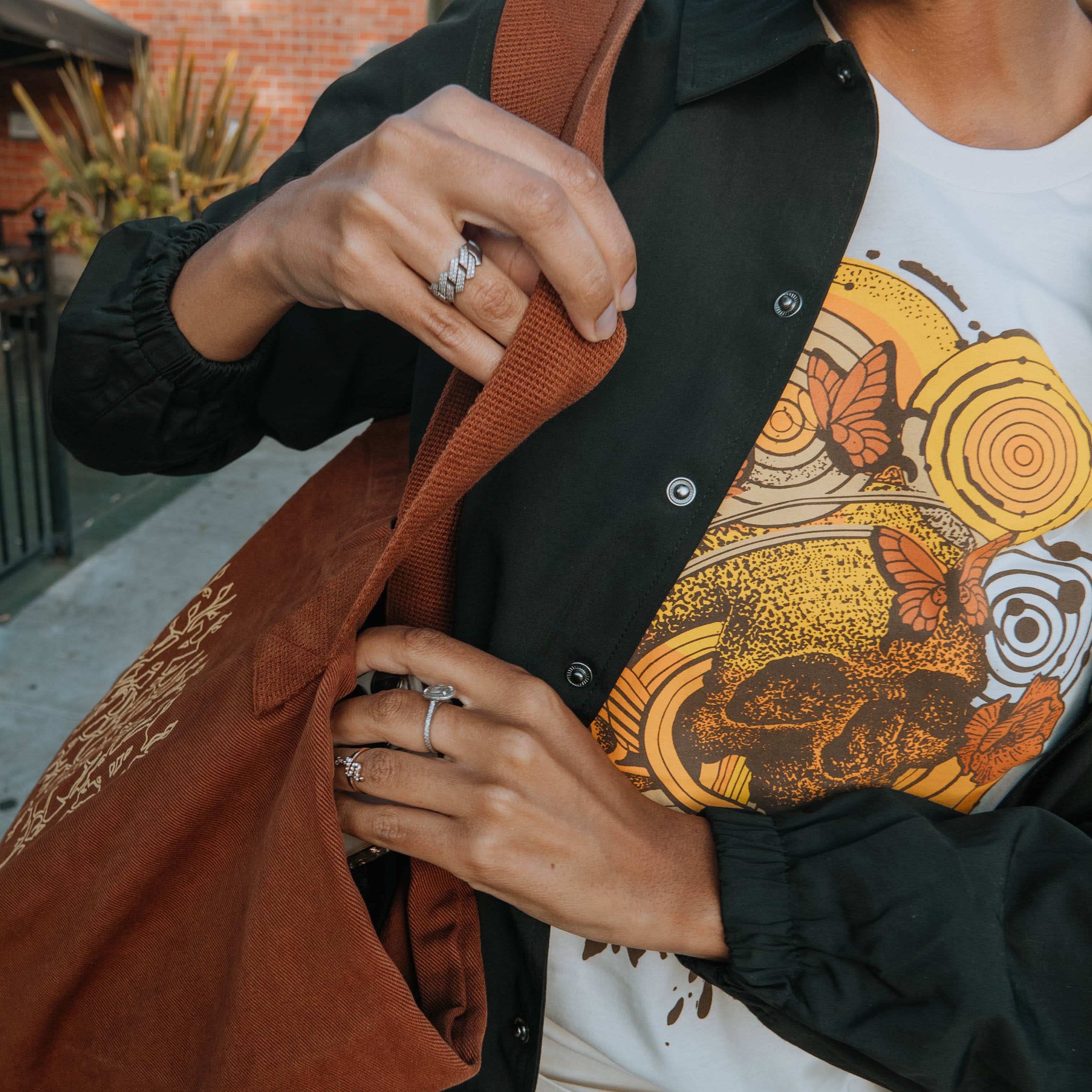Woman (but you can't see her face) wearing Rebirth tee in natural with skull plus flowers and geometric designs in oranges and yellows, black jacket, and brown tote bag, hands reaching inside bag.