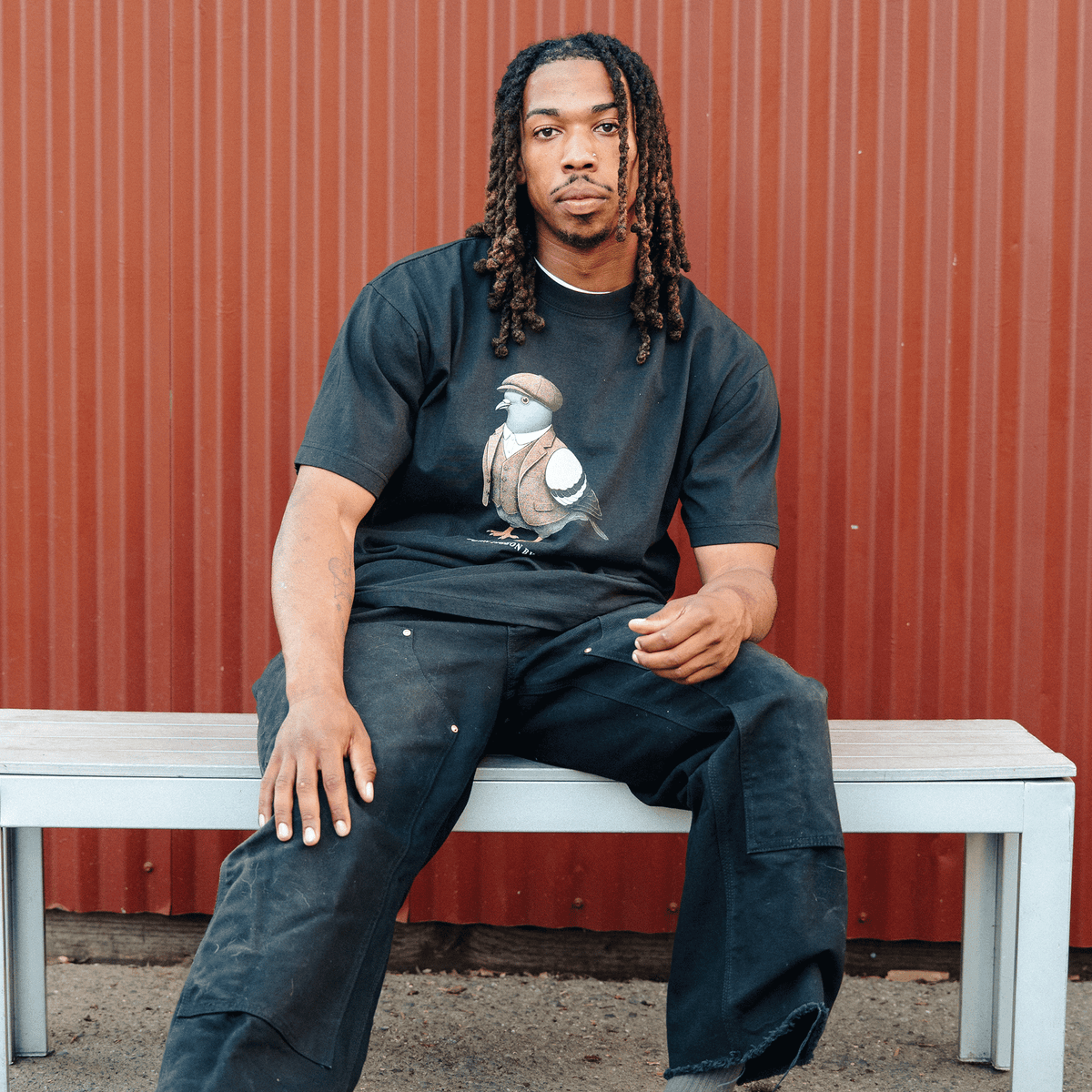 A man with shoulder-length dreadlocks wears the black Oaklandish Dapper Tee featuring a graphic of a pigeon in a tweed vest and flat cap, sitting on a white bench against a corrugated red metal wall in loose black work pants.
