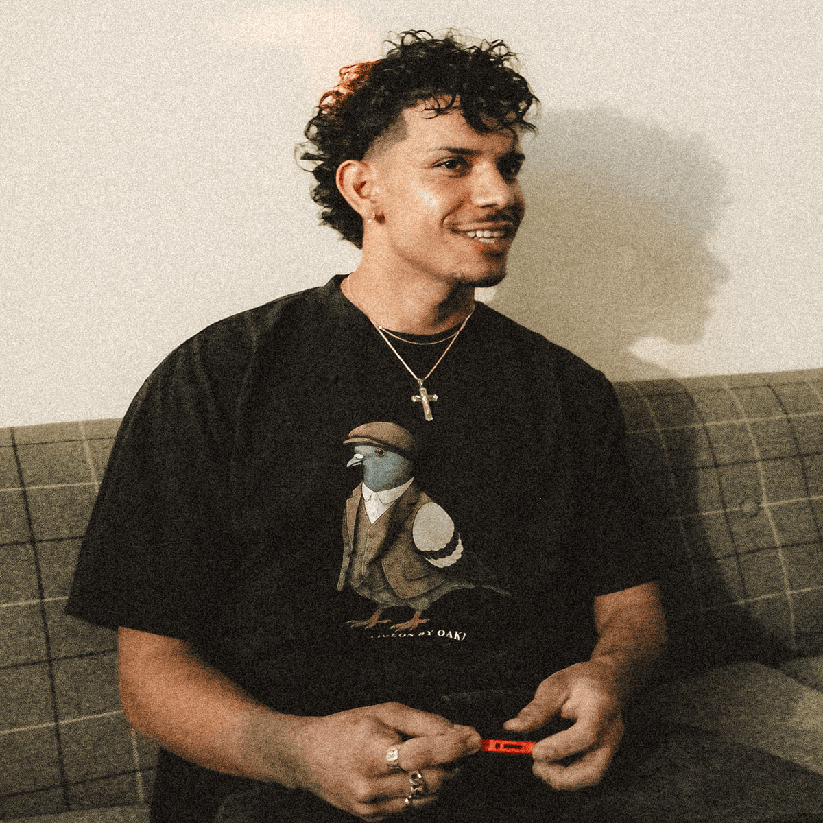 A young man with curly hair and a goatee sits on a couch wearing an Oaklandish Dapper Tee featuring a pigeon in a vest and flat cap, accessorized with a silver cross necklace and rings.