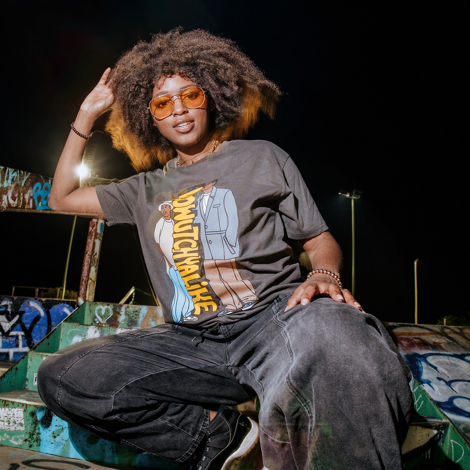 A person with curly hair and orange-tinted glasses confidently poses on graffiti-covered steps, channeling Digital Underground's 90s vibes. Their "Doowutchyalike" tee by Oaklandish, featuring an illustration and text, pairs with dark pants and a bracelet against a night sky backdrop.