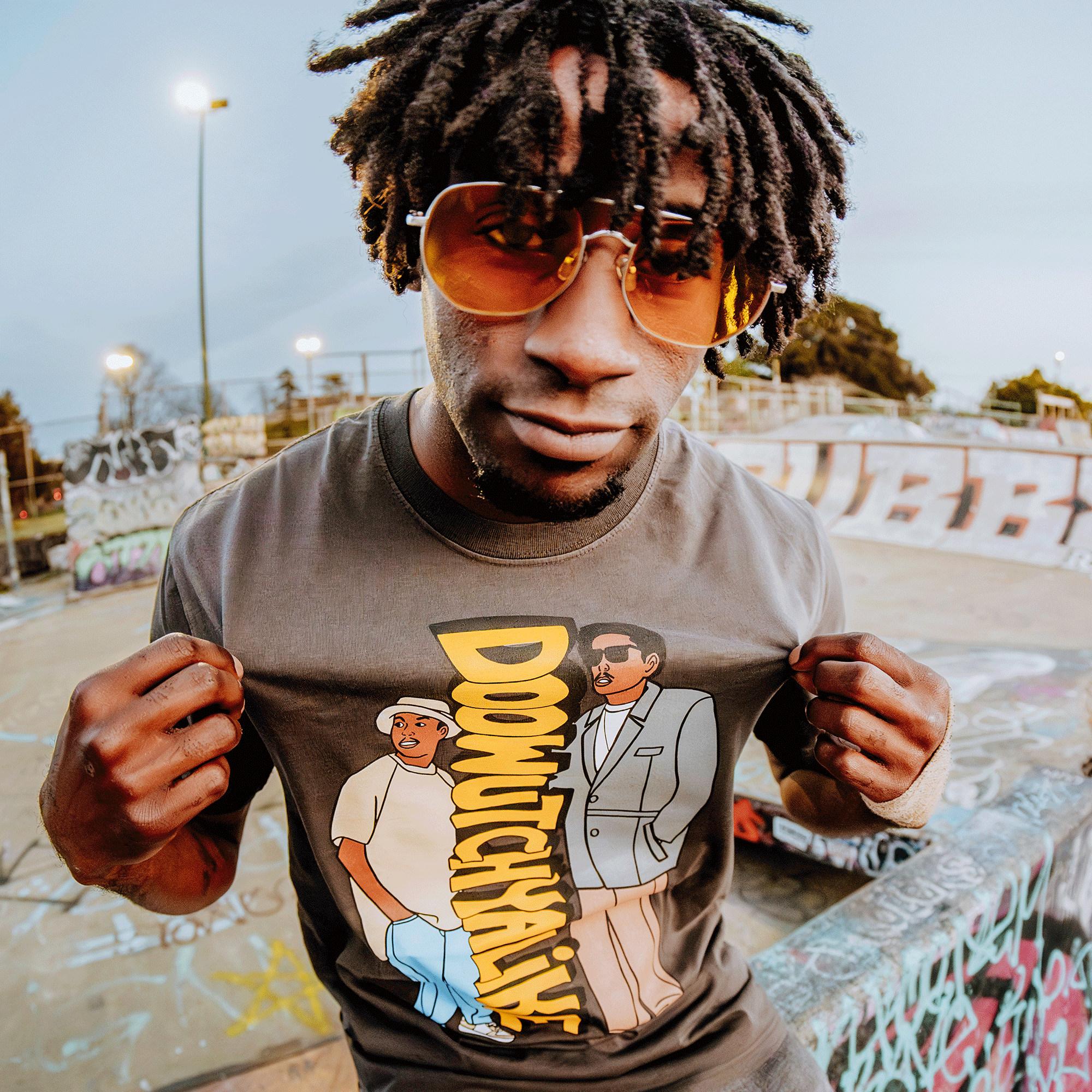 Wearing yellow sunglasses and the Doowutchyalike Tee by Oaklandish, a man with dreadlocks embodies 90s style at a graffiti-covered skatepark. He poses against vibrant street art under an overcast sky, celebrating his shirt's Digital Underground-inspired design.