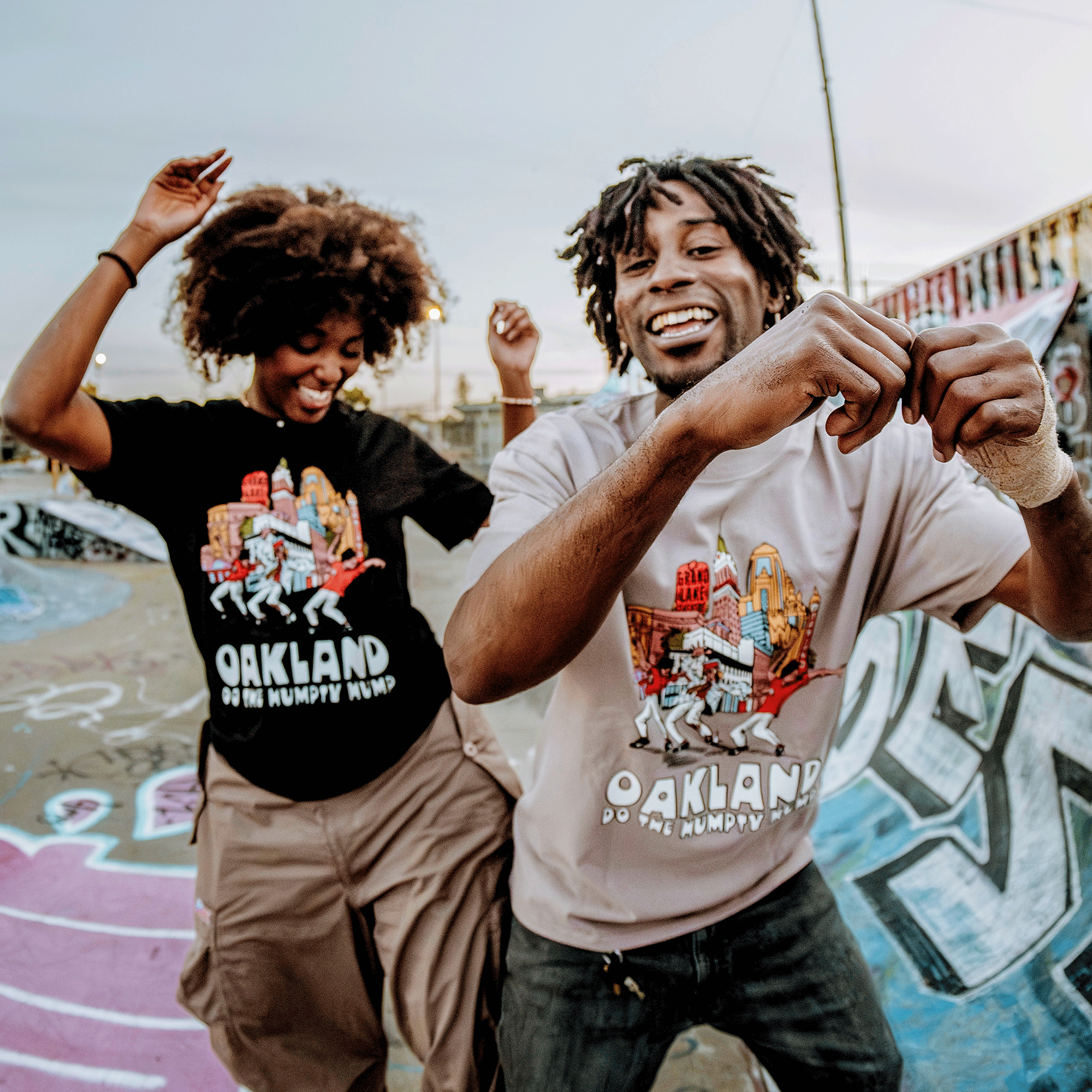 In a graffiti-covered skate park, two people dance joyfully to Digital Underground's "Humpty Hump." Both wear Oaklandish T-shirts: one in black with beige pants and the other in light grey with jeans.