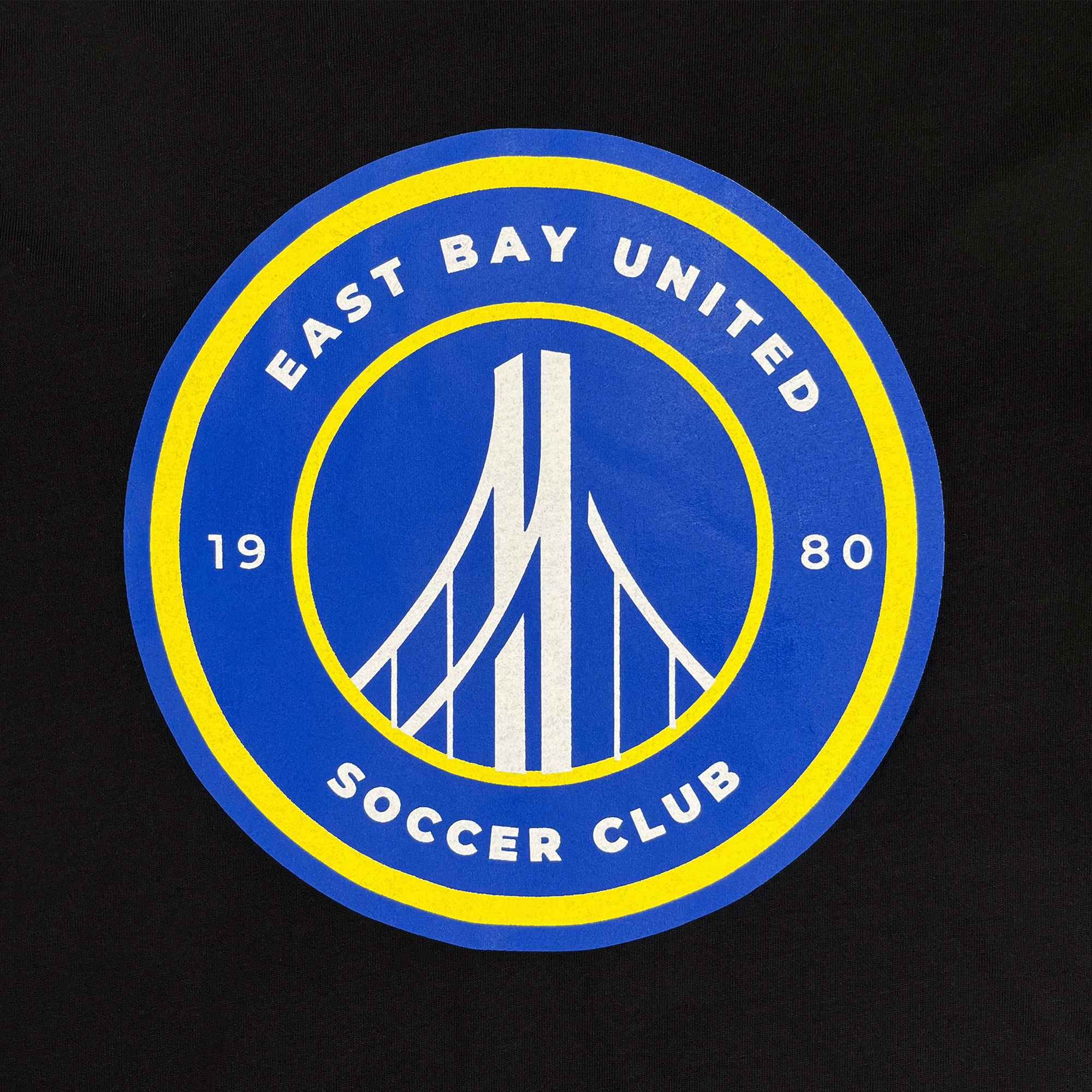 Close-up of round blue, white, and yellow East Bay United Soccer Club 1980 logo on the front chest of black t-shirt.