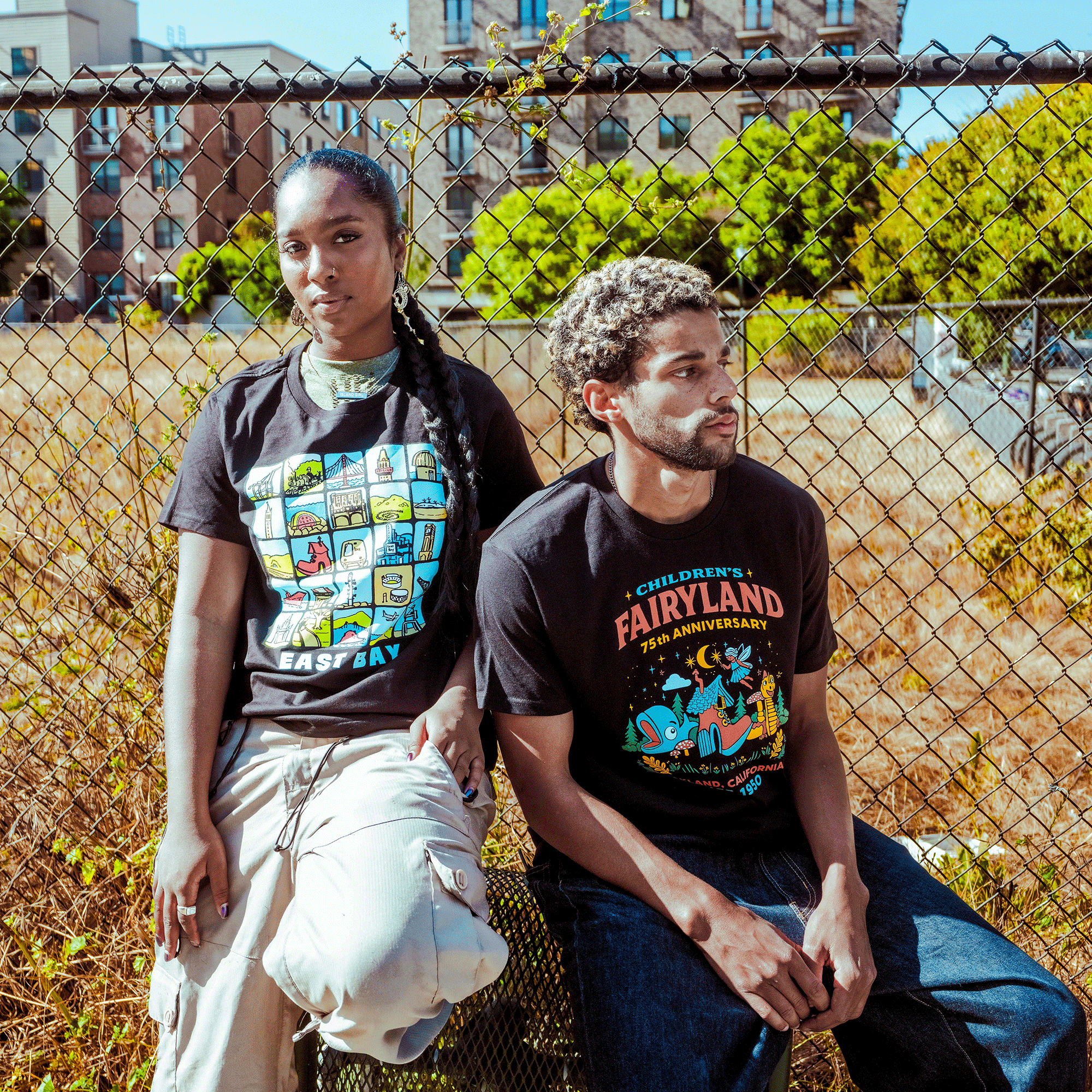 A young man with short curls and a beard sits by an urban chain-link fence, wearing jeans and the Oaklandish East Bay Landmarks Tee by a local artist. Next to him, a woman sports long braids, light pants, and a black cartoon graphic tee.