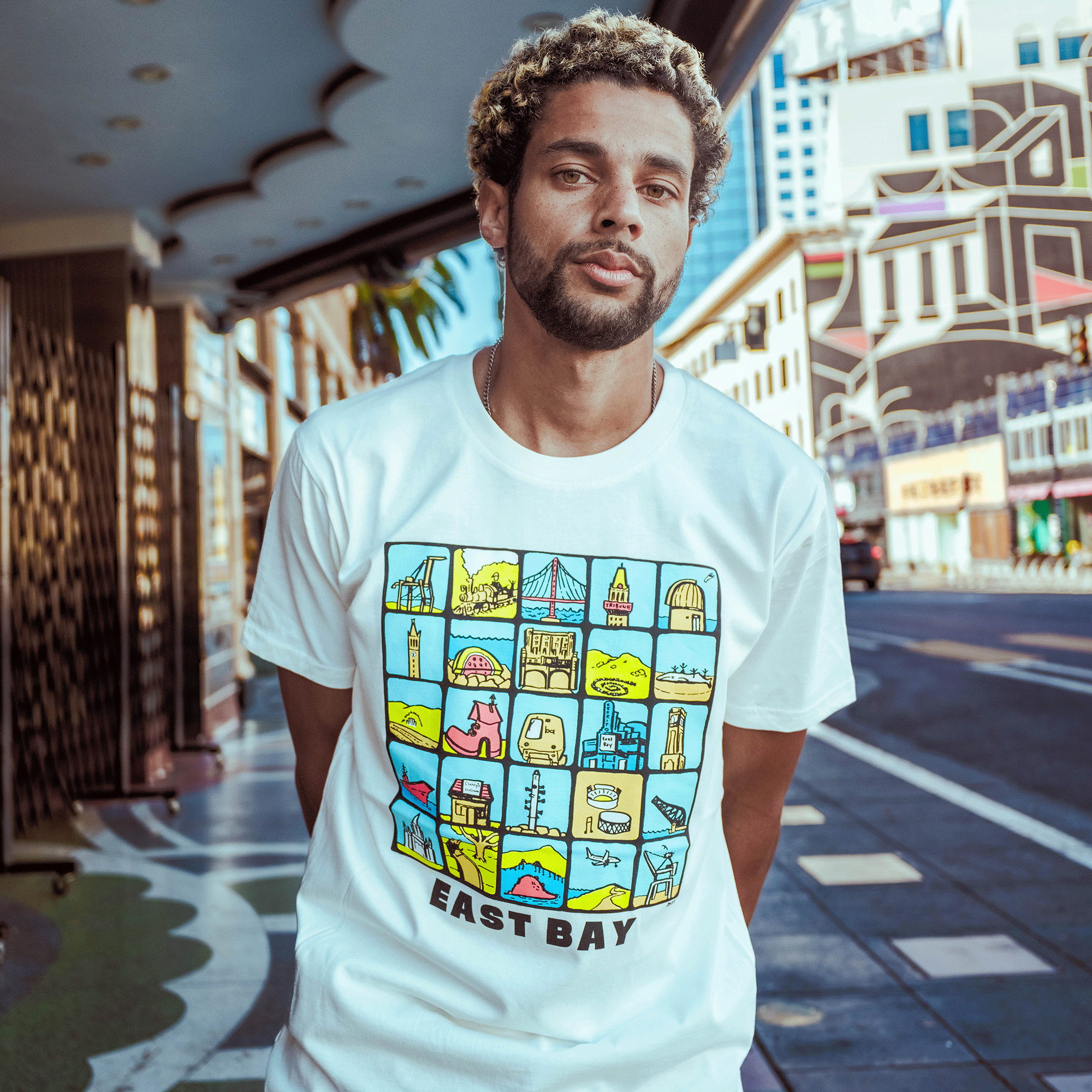 A bearded man with short curly hair stands on a city sidewalk, wearing the Oaklandish East Bay Landmarks Tee featuring colorful cartoon Bay Area landmarks. Buildings and palm trees can be seen in the background.