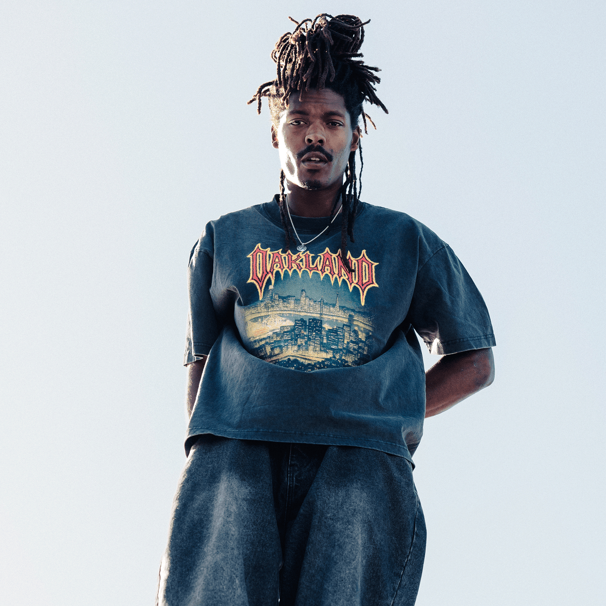 A man with long dreadlocks looks down at the camera, wearing an Oaklandish Ember Tee in charcoal grey with a stylized “OAKLAND” skyline graphic. Framed against a pale sky, he wears dark denim pants and a thin silver necklace.