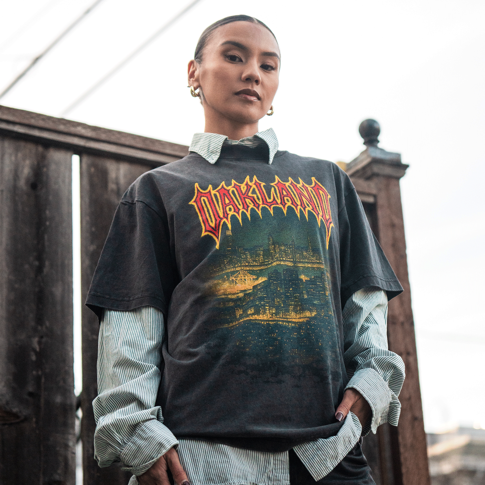 A confident man stands by a wooden fence, wearing the black Ember Tee by Oaklandish (100% cotton, classic fit) over a striped button-up. He has slicked-back hair, gold hoop earrings, a serious look, and one hand in his pocket.