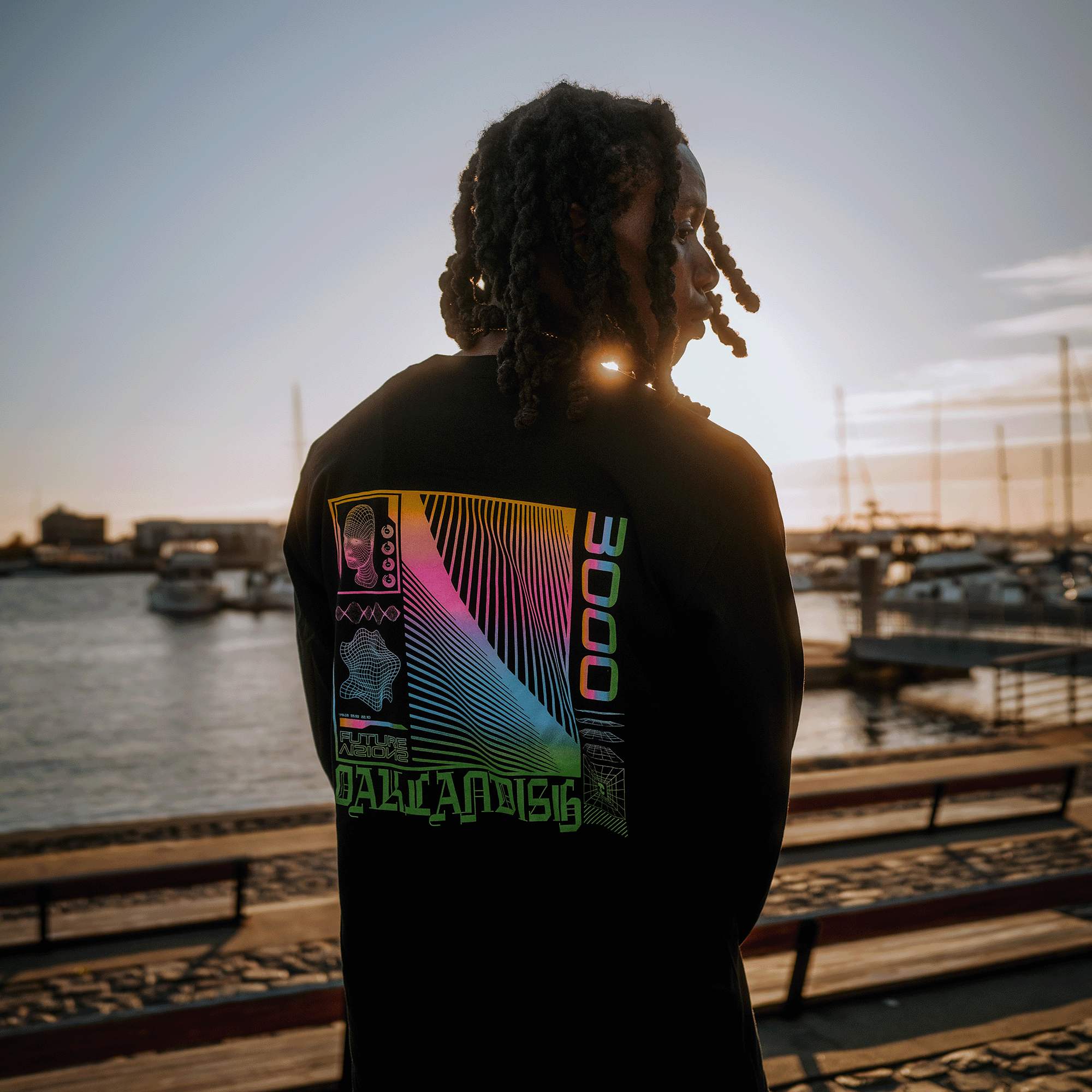 Backside view of model wearing black long-sleeve t-shirt with neon pink, yellow, blue, and green futuristic graphic with OAKLANDISH wordmark.
