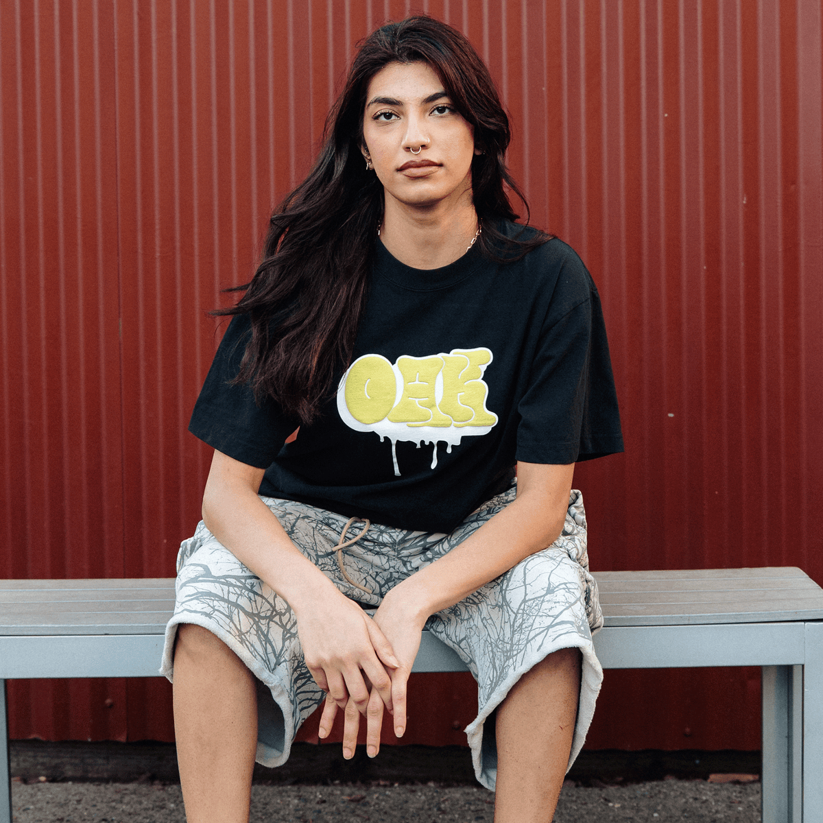 A person with long dark hair and a septum piercing sits on a bench against a red corrugated metal wall, wearing an Oaklandish Graff OAK Tee featuring yellow graffiti-style letters with white drip accents and light-colored shorts with an intricate branch-like pattern.