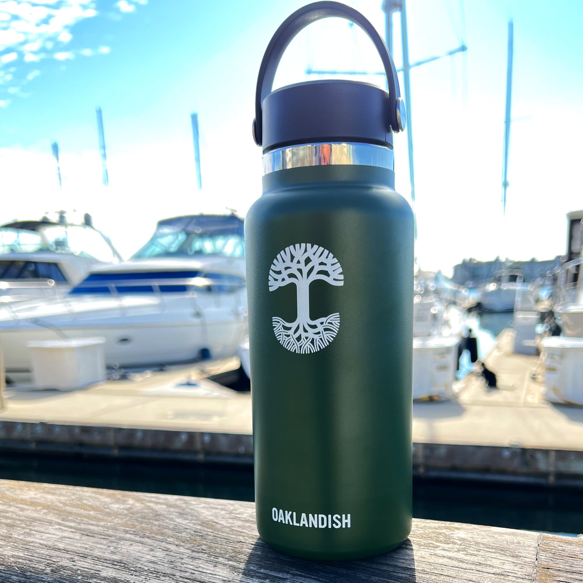 An Oaklandish Canteen, featuring a dark green, double-wall insulated design and a convenient handle, is placed on a wooden surface near a marina with several docked boats. The canteen showcases a white graphic of a stylized tree and the word "OAKLANDISH" in white text beneath it. The scene captures a sunny day under a clear, blue sky.