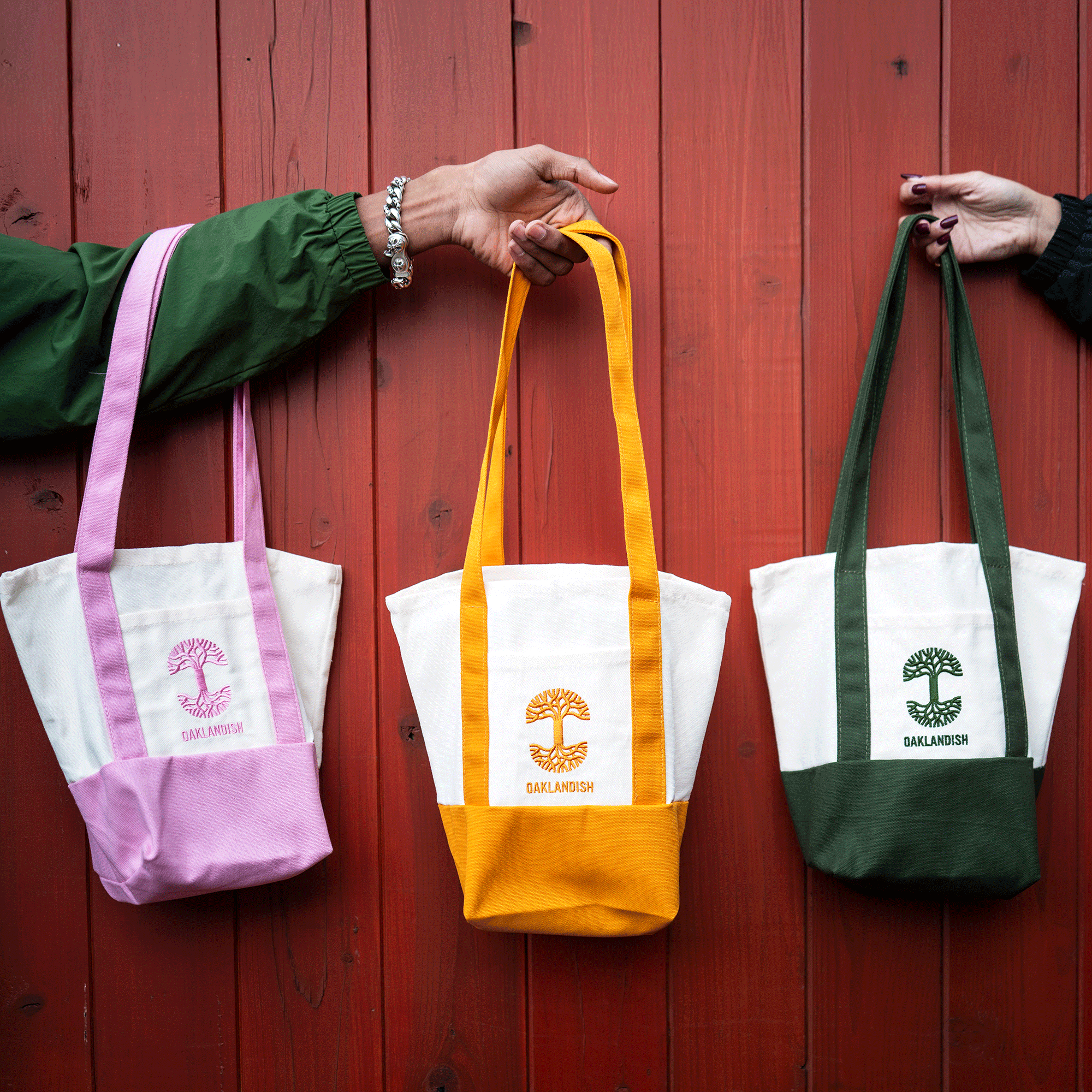 Two hands hold three Oaklandish Mini Grocery Totes by Oaklandish in front of a red wooden wall. The white canvas bags have colored straps and bases—pink, yellow, and dark green—and feature an embroidered “OATLANDISH” tree logo on the front.