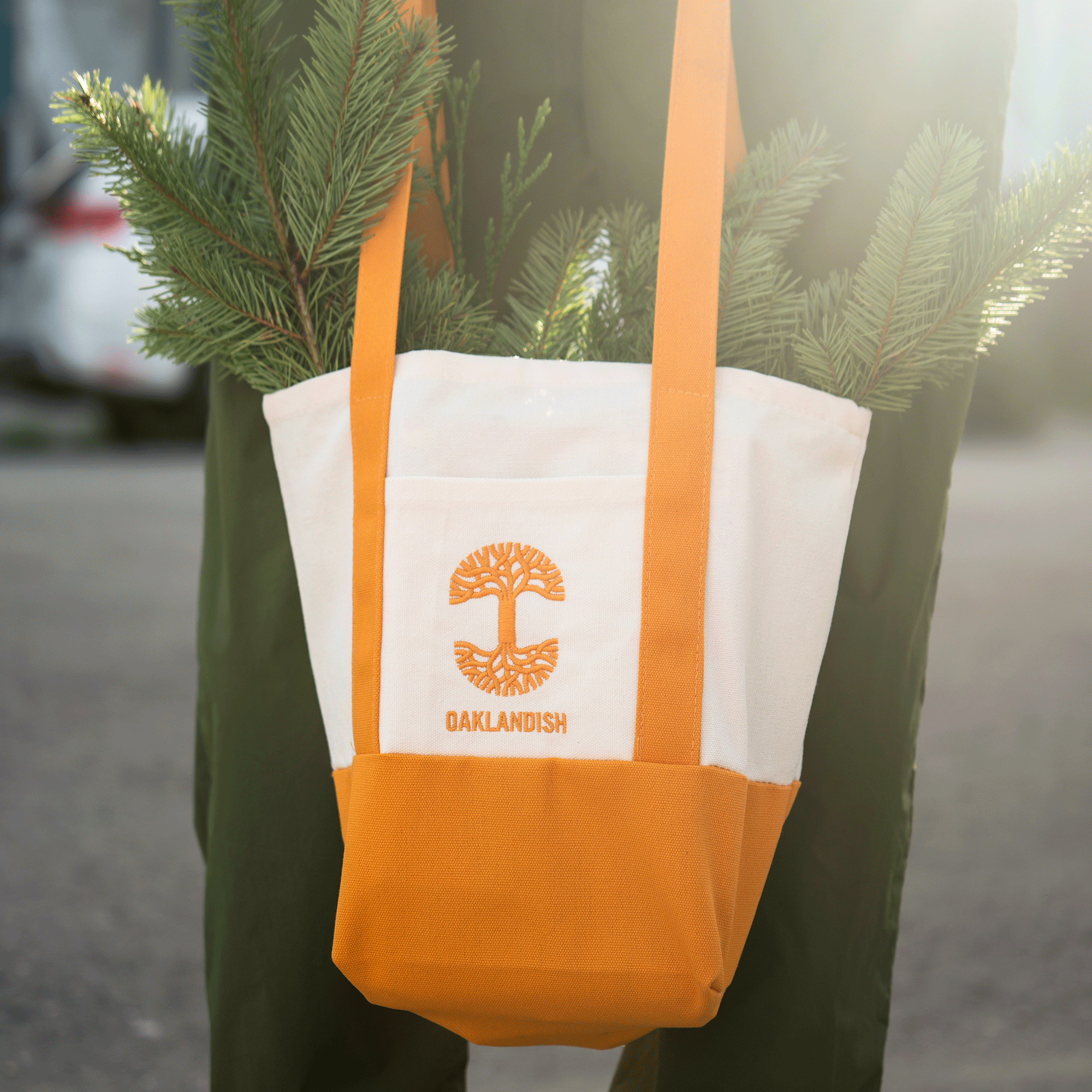 A person holds the Oaklandish Mini Grocery Tote by Oaklandish, a white and orange bag with embroidered logo and tree, filled with pine branches. They wear dark green pants and stand outdoors on sunlit pavement.