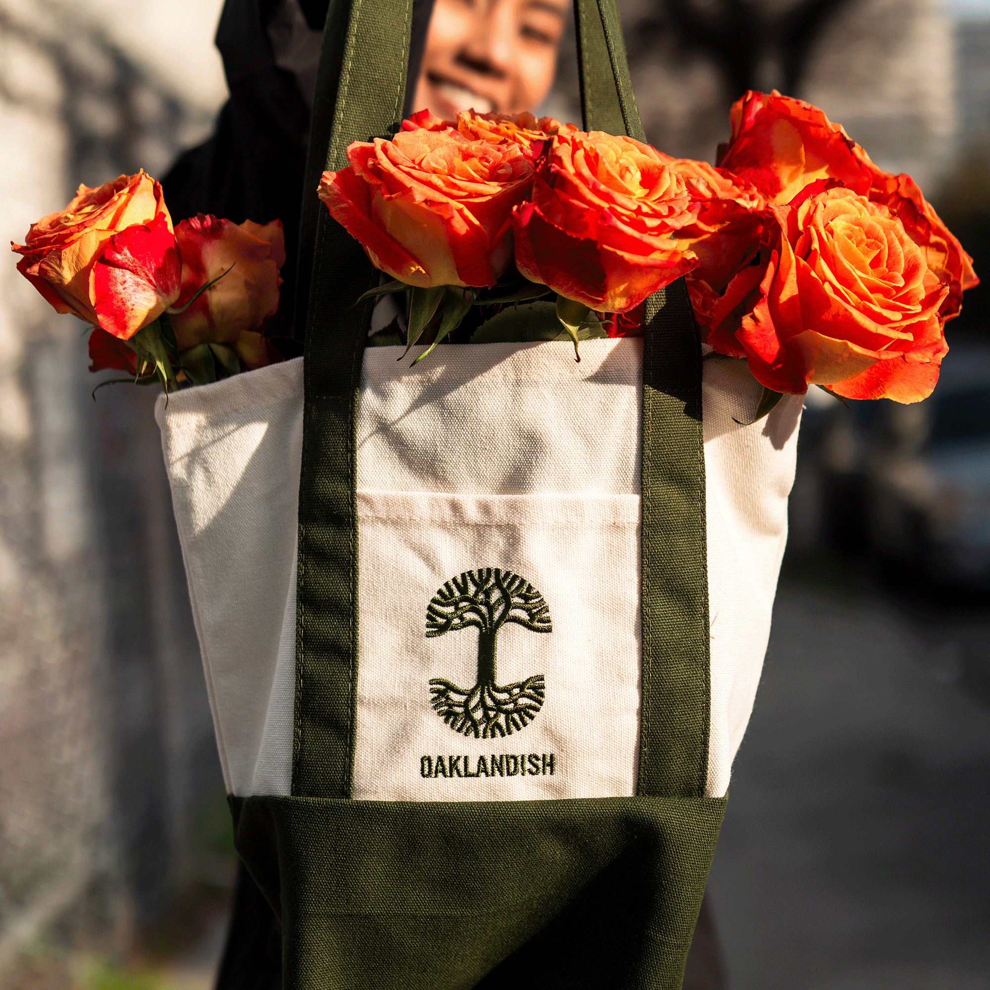 A smiling person holds the Oaklandish Mini Grocery Tote by Oaklandish, with green straps and logo, filled with vibrant orange roses. The outdoor background is slightly blurred, showing a fence and a car.