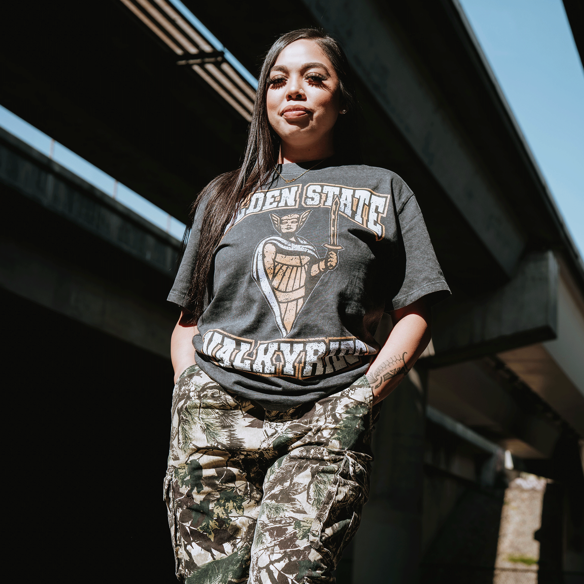 A woman with long dark hair stands confidently under a sunlit bridge, wearing the GSV Golden State Valkyries Guardian Stone Wash Heavy Tee and camouflage pants. Blue sky and shadows appear in the background.