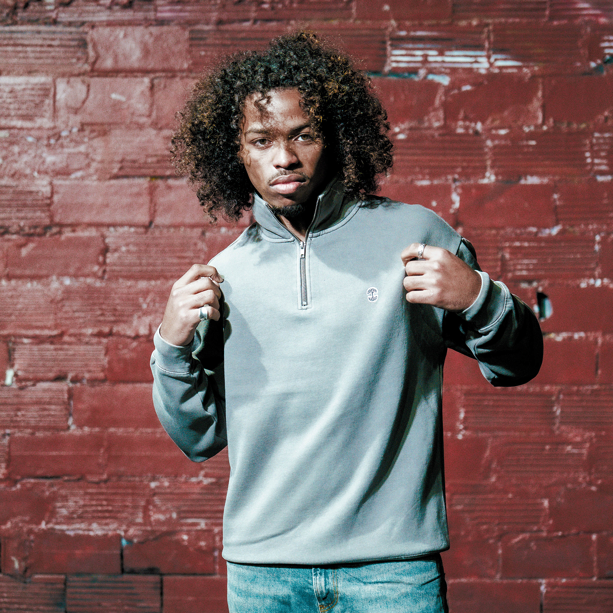 A young man with curly hair stands before a red brick wall, confidently holding the collar of his Oaklandish Hojicha 1/4 Zip pullover while wearing blue jeans.