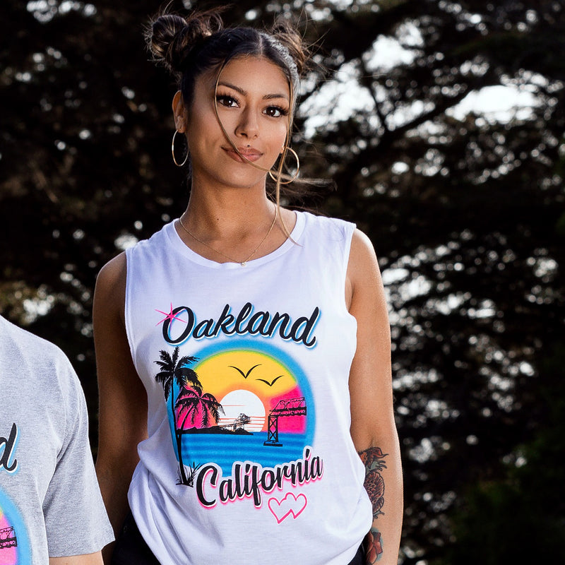 Model outdoors wearing white tank top with multicolor bright ink colors coastal bay scene with palm trees and bridges and hearts, text spelling Oakland, California.
