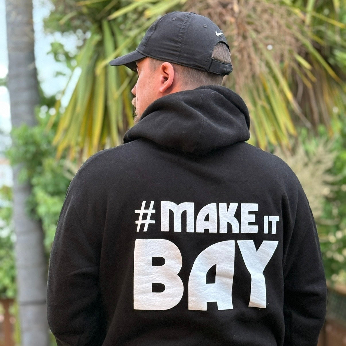 A person in a black Oaklandish Make It Bay Hoodie faces away outdoors, "#MAKE IT BAY" boldly displayed on the back. Lush green palms set the scene, highlighting its nod to Bay Area creatives and local collaboration.