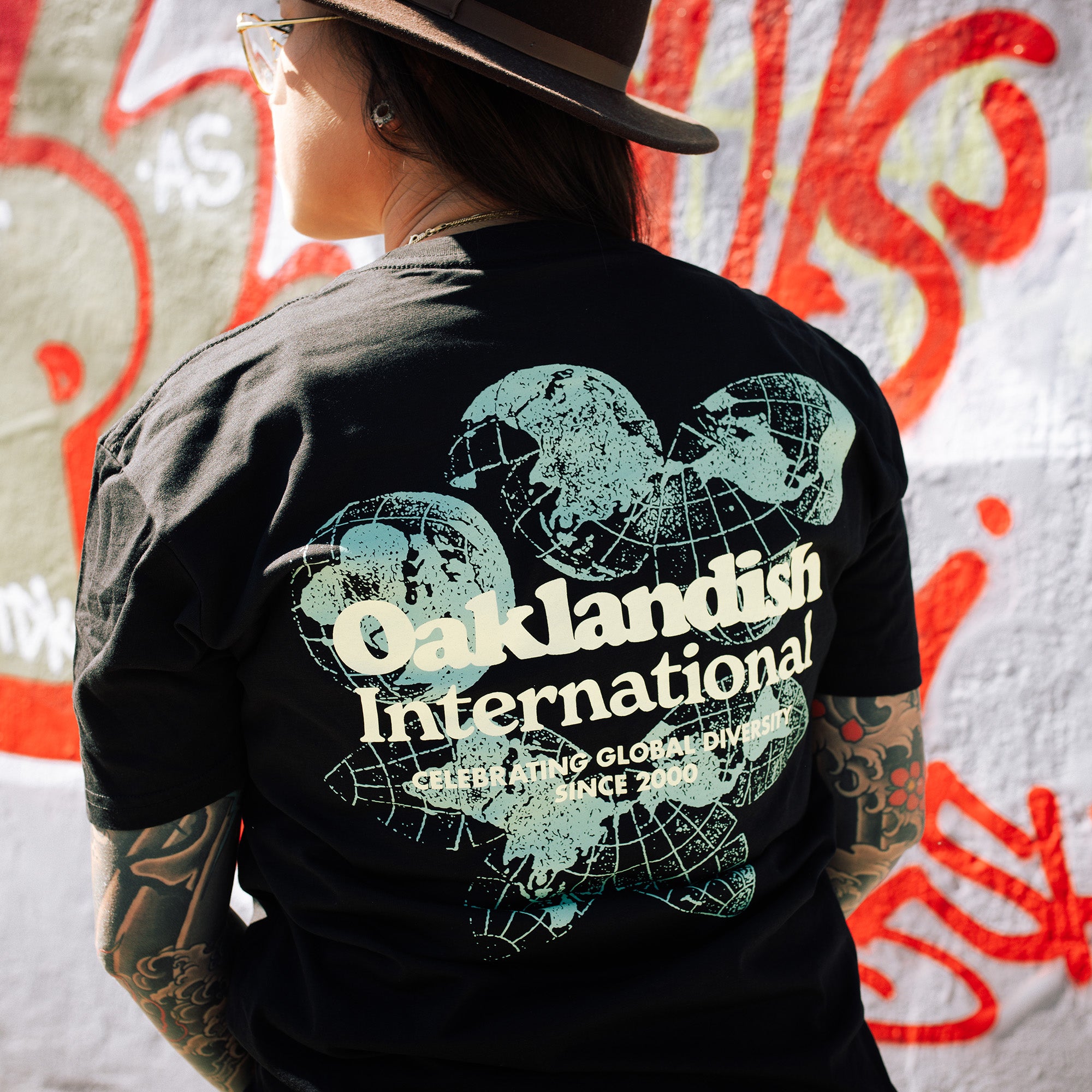 Female model showing off backside of a black t-shirt featuring a green and yellow Oakland International graphic with a caption celebrating global diversity since 2000.
