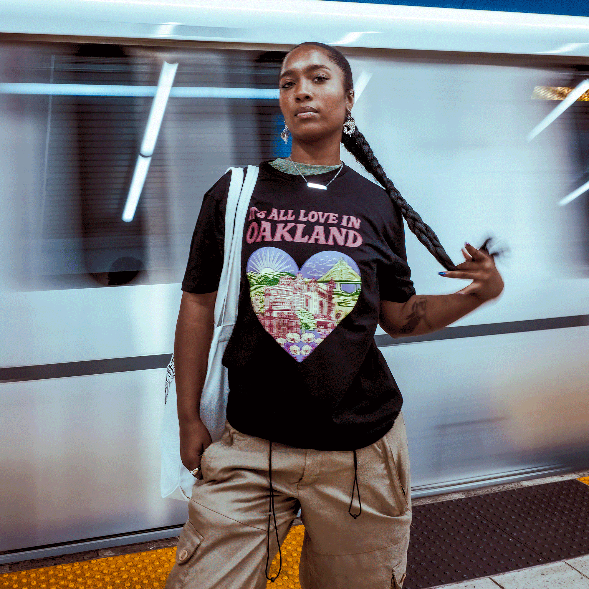 A confident person with long braids stands on a subway platform in tan cargo pants and the Oaklandish "It's All Love in Oakland Tee," made from 100% cotton and featuring a colorful heart graphic, as a train blurs past behind them.