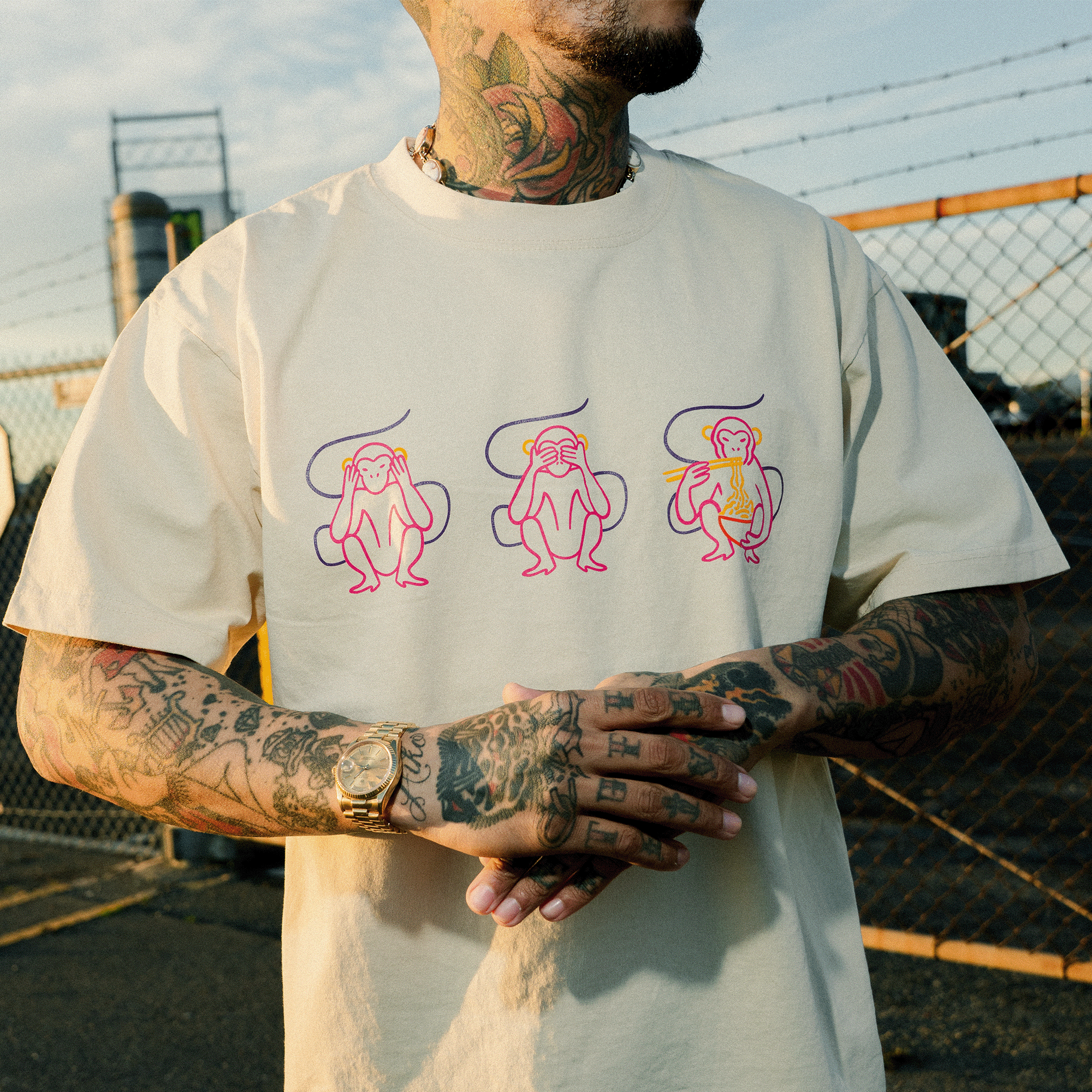 A tattooed man stands outdoors, fingers interlaced, sporting the Oaklandish Jo's Modern Thai X Oaklandish Tee—a playful heavy-weight shirt featuring cartoon outlines of three monkeys: see no evil, hear no evil, speak no evil.