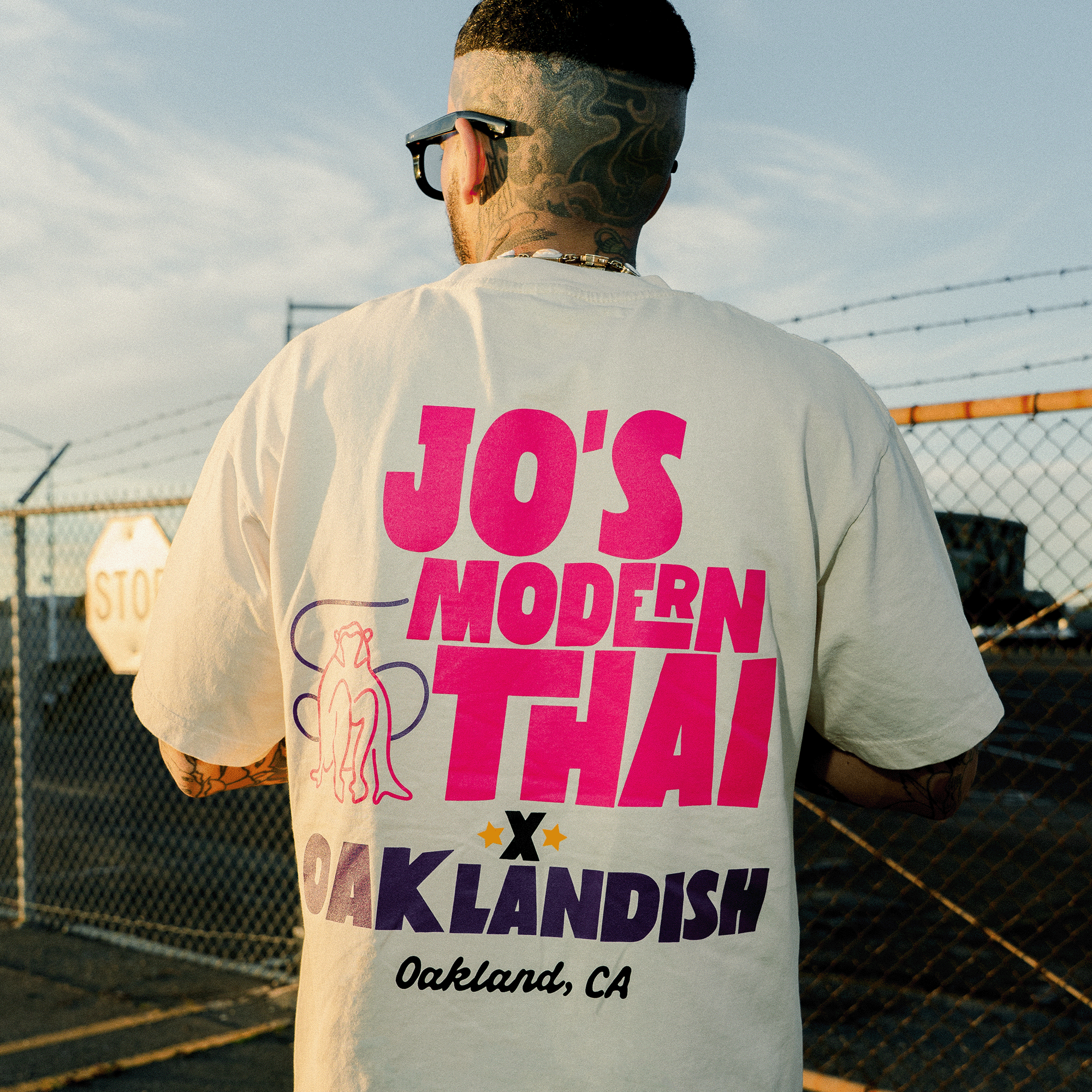 A tattooed person stands outdoors in the Oaklandish Jo's Modern Thai X Oaklandish Tee, showcasing bold collaborative text and playful graphics. A fence and stop sign in the background capture the Oakland, CA vibe.