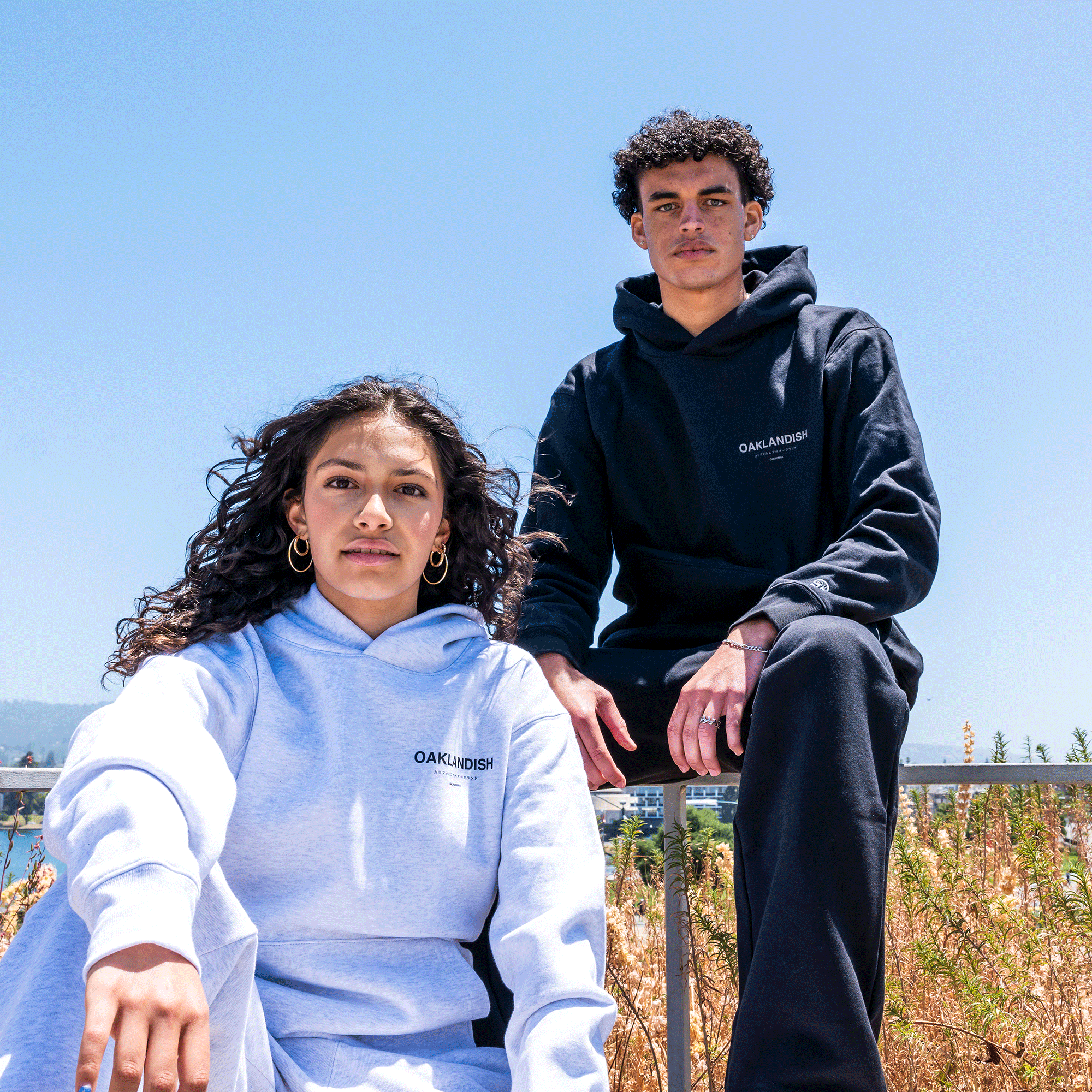 A young woman and man stand outdoors wearing Modern Type Hoodies from the brand "Oaklandish." The woman wears a light gray organic cotton hoodie emblazoned with the brand name in black, while the man sports a black hoodie featuring the brand name in white. They are surrounded by a bright blue sky and tall grass.