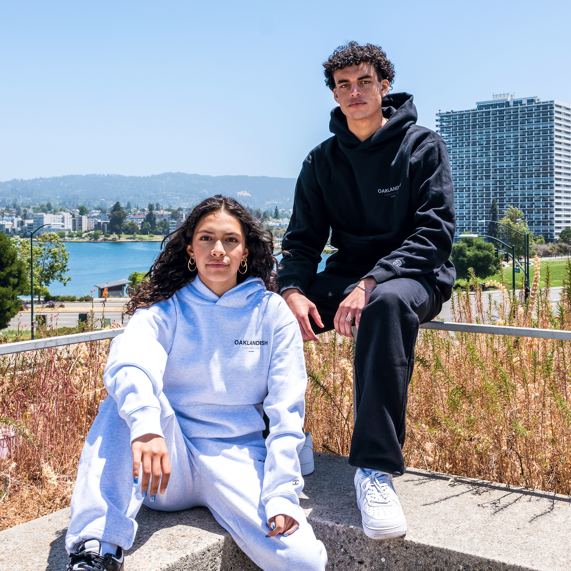 Two individuals, one woman and one man, are posing outside in casual attire. The woman is seated on a concrete ledge wearing light gray Oaklandish Modern Type Sweatpants and an organic cotton hoodie. The man stands next to her in black Oaklandish Modern Type Sweatpants and a matching hoodie. Both hoodies display a small logo. An urban park and buildings are visible in the background.