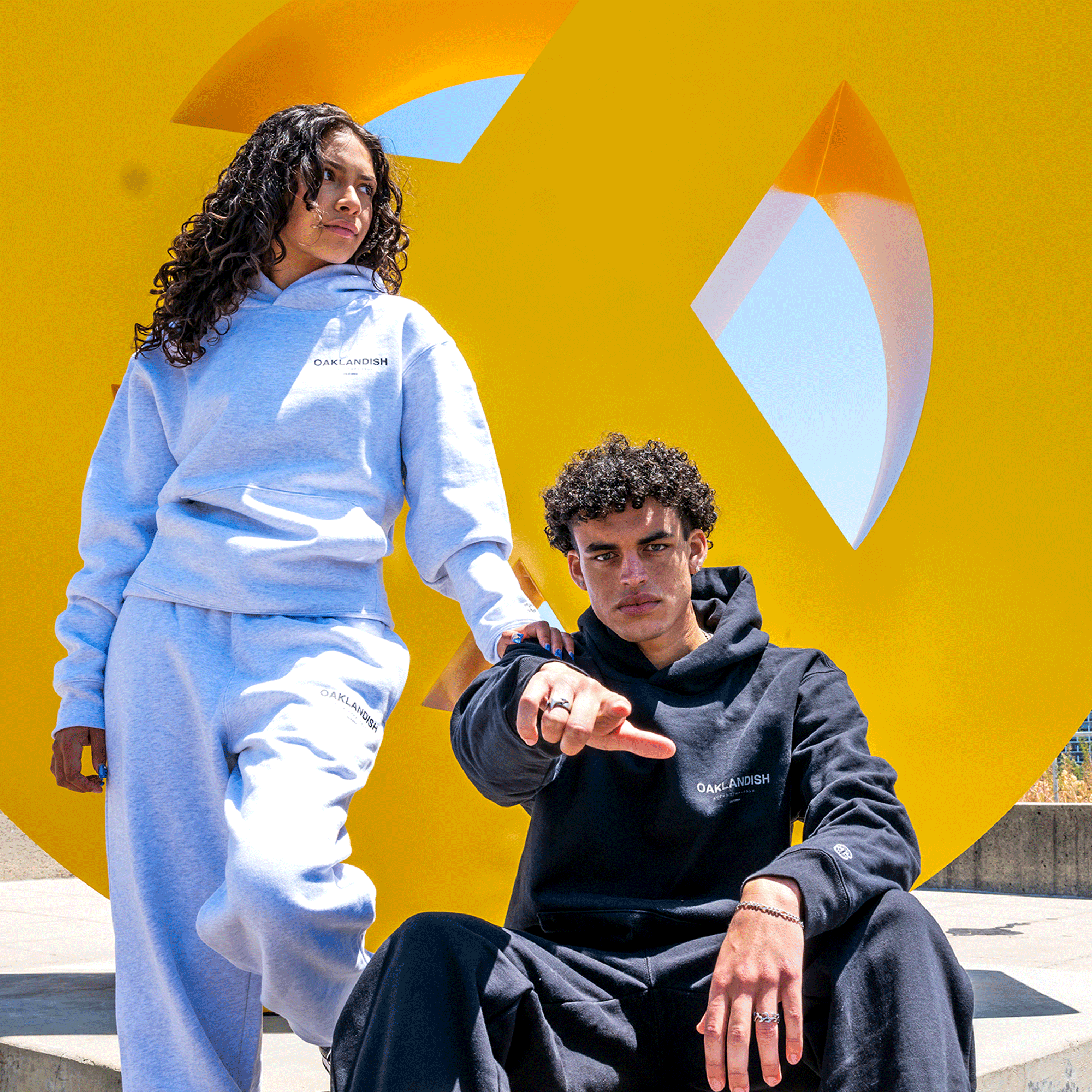 Two individuals in casual wear. One stands in a light blue tracksuit with "Oak and Ashen" printed on the chest, while the other sits in black Modern Type Sweatpants and a matching hoodie from the unisex brand Oaklandish, pointing forward. They pose against a bright yellow abstract sculpture under clear blue skies.