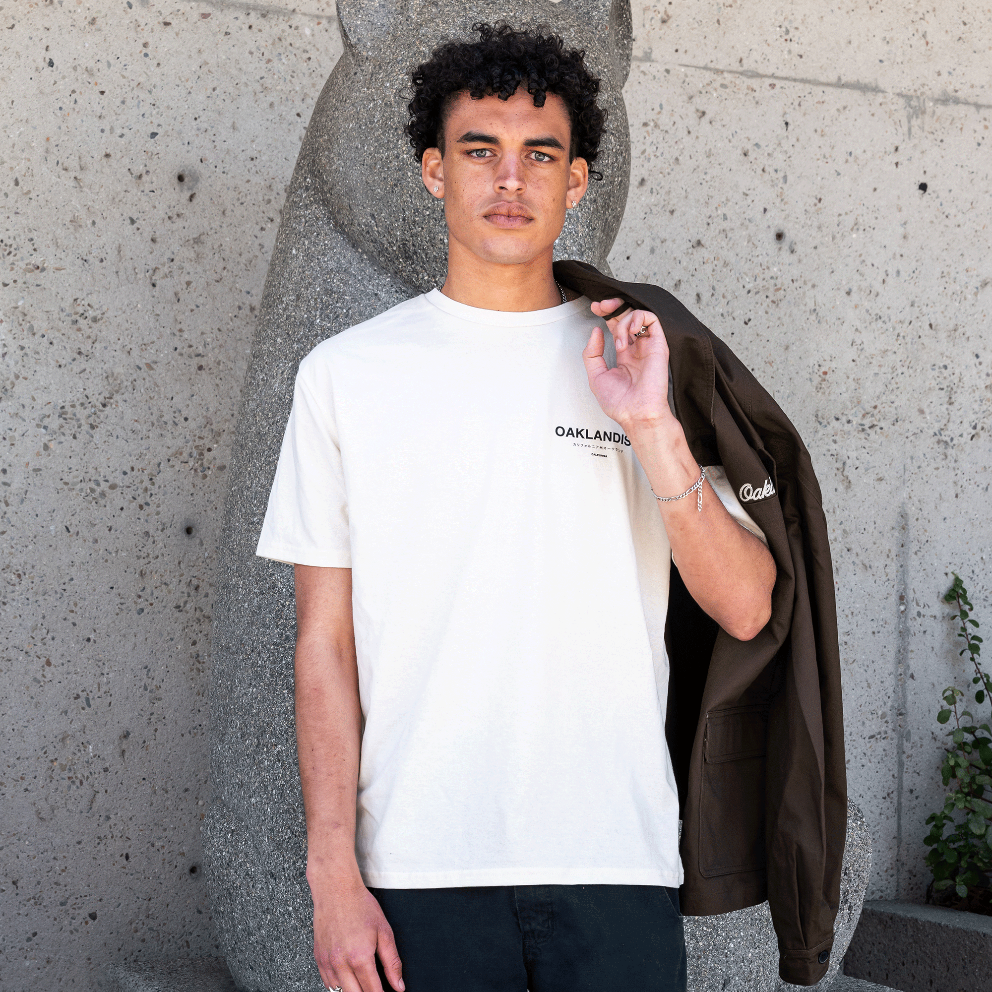 A young man with curly hair stands in front of a textured concrete wall and stone sculpture. He is wearing the Oaklandish Modern Type Tee from the men's collection paired with black pants. Holding a brown jacket slung over his shoulder, he looks directly at the camera. There is a small plant on the right.