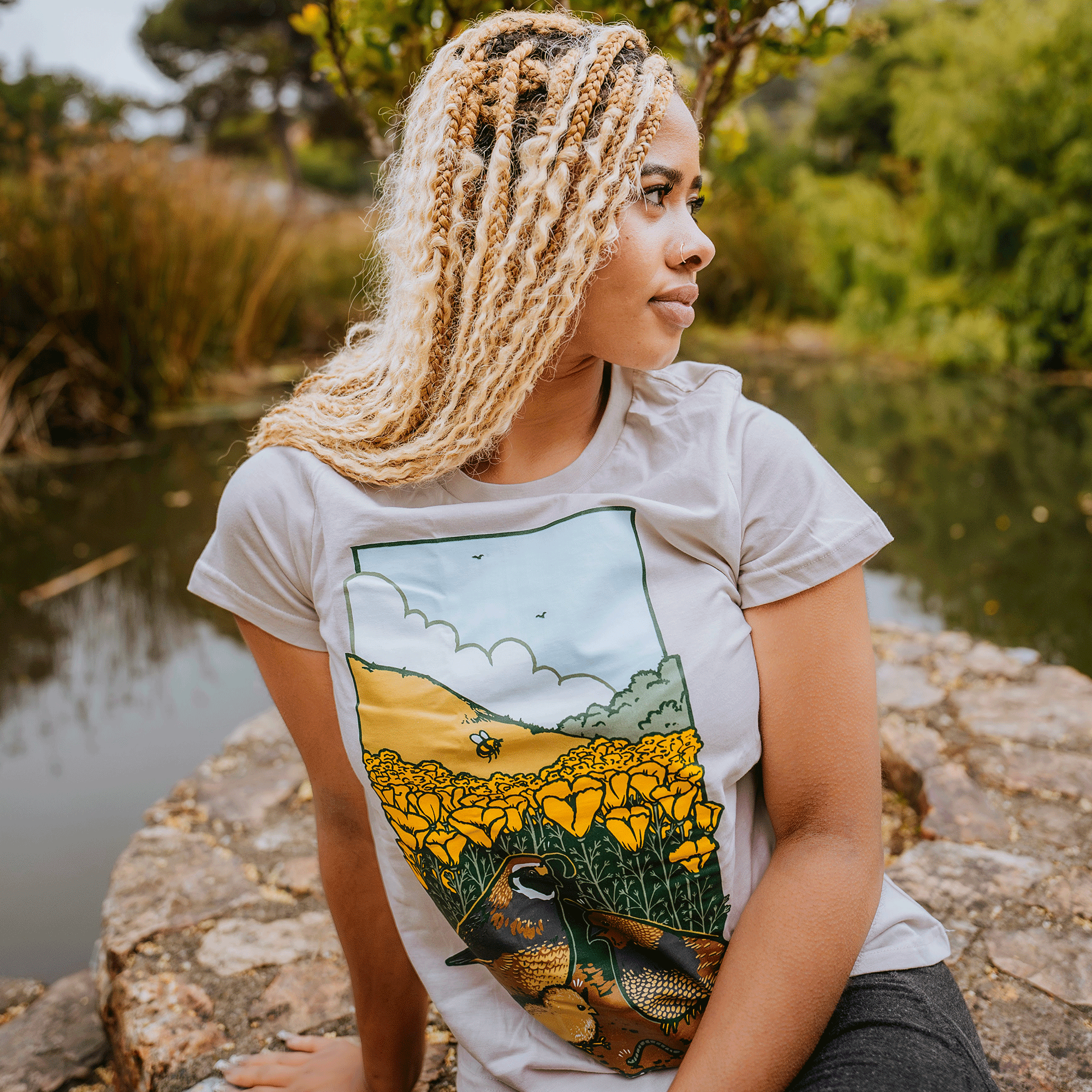 A woman with blonde braids sits by a pond on a stone ledge, gazing to the side. She's wearing the Women's Quail Family Tee from Other, which is 100% cotton and fits perfectly. The tee features an illustration of yellow flowers and quails, complementing the serene greenery around her.